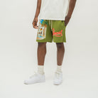 A person modeling olive green drawstring shorts with a colorful tropical beach graphic on the left leg and red 'Almost Someday' text on the right leg, paired with a white t-shirt, white socks, and white sneakers against a plain white studio background.