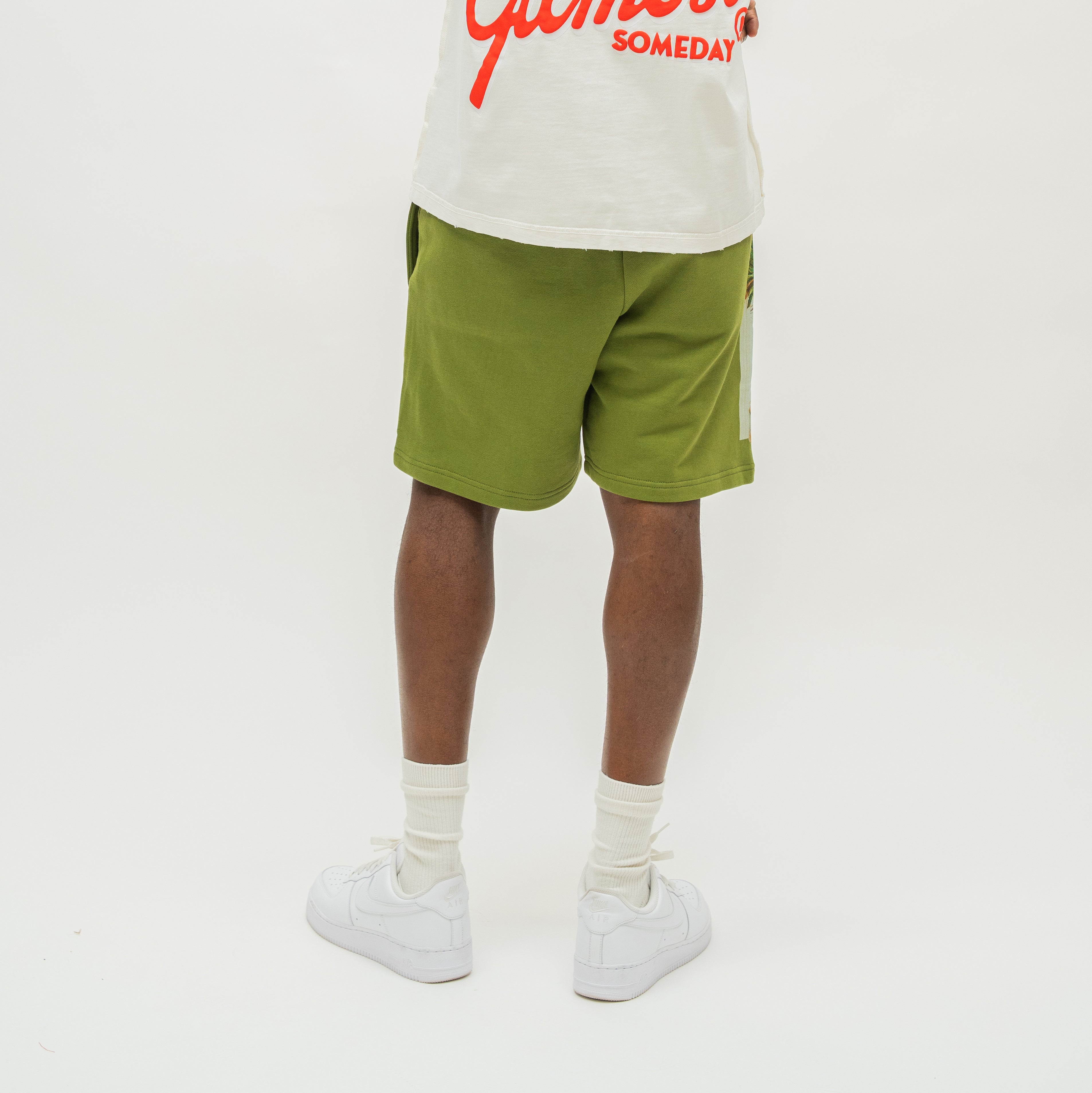 A model from the waist down wearing olive green athletic shorts, a cream graphic t-shirt, white socks, and white sneakers, standing against a plain white studio background.