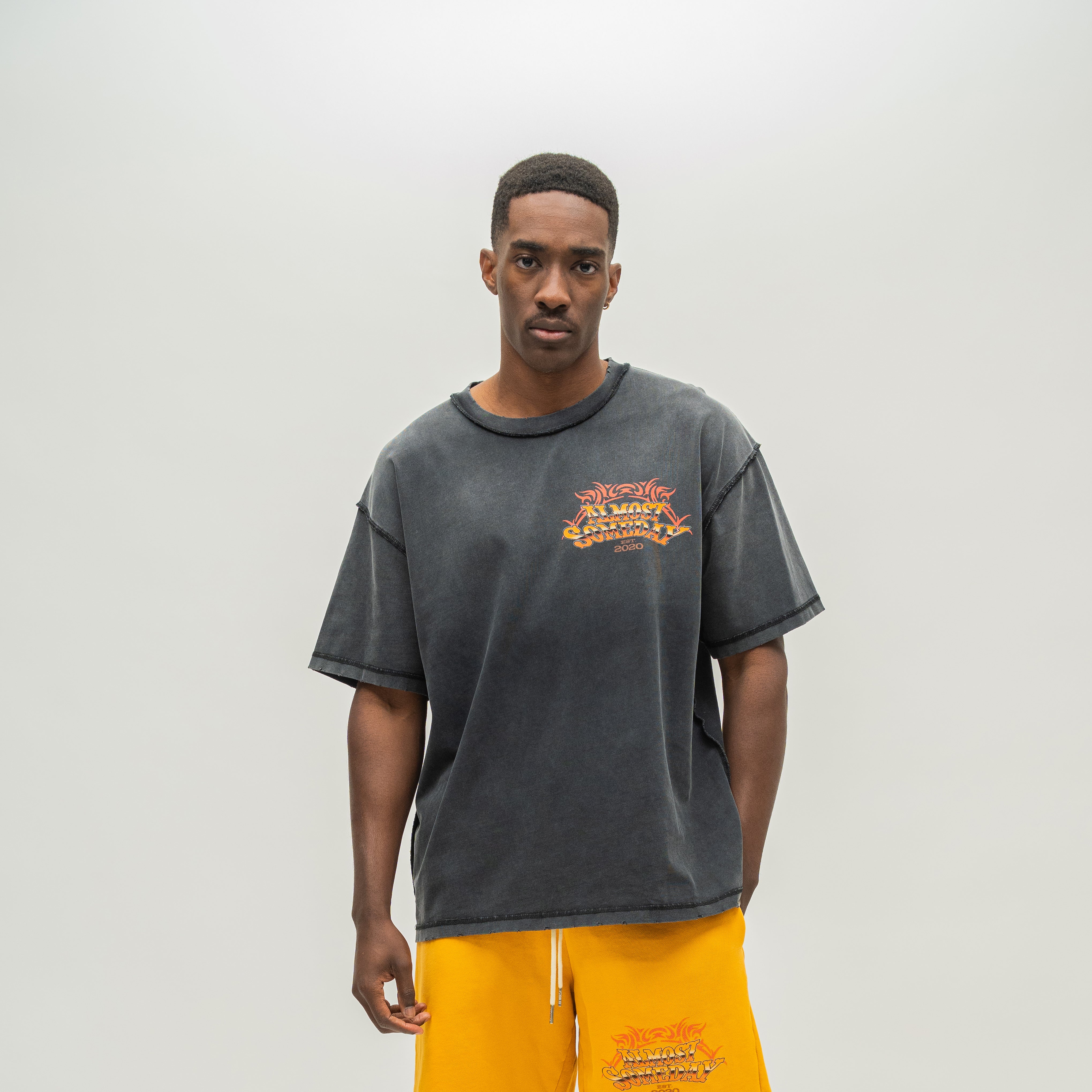 A man modeling an oversized faded black acid-wash t-shirt with a distressed orange graphic on the left chest, paired with bright yellow sweatpants, against a plain light gray background.