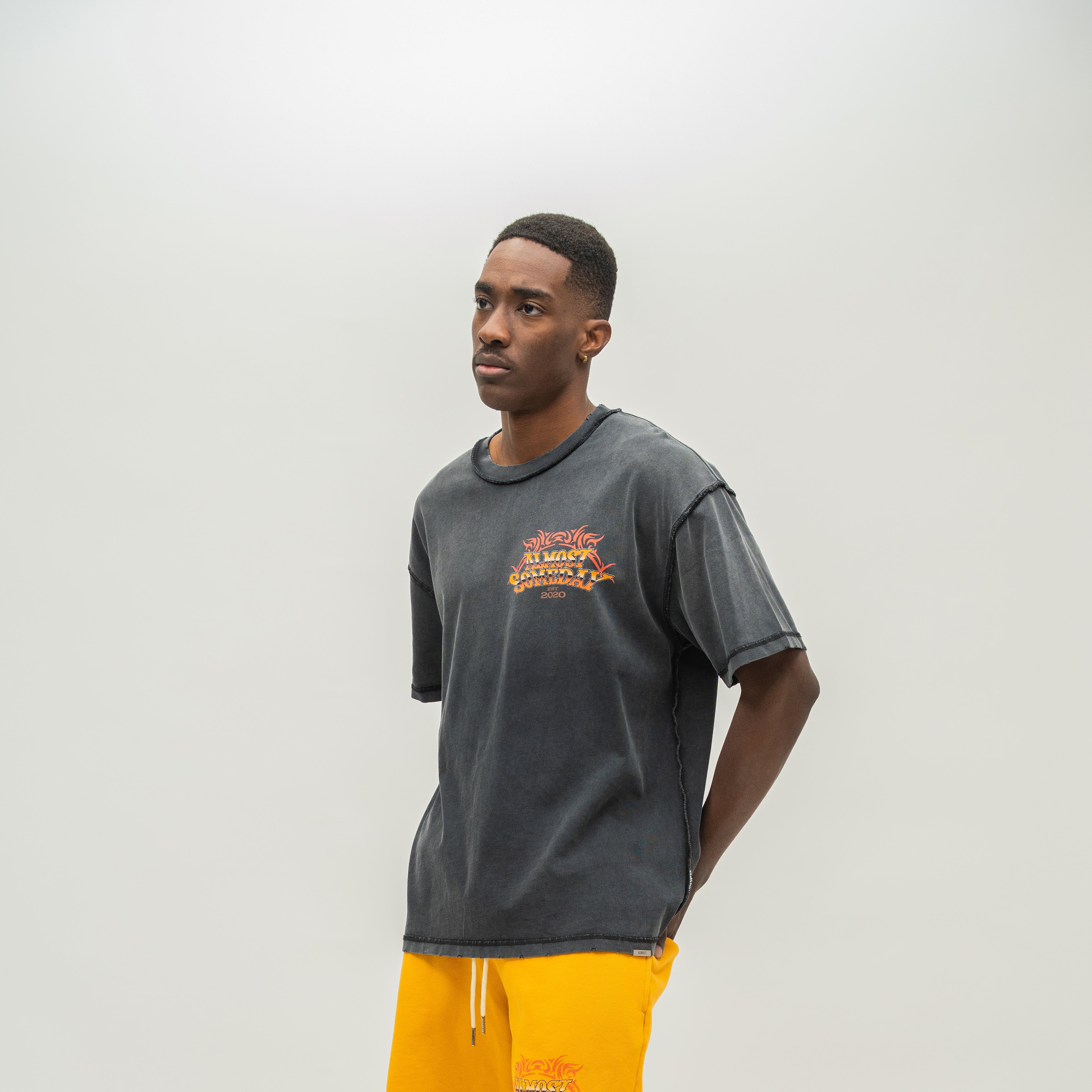 A man wearing a washed black t-shirt with a yellow and red graphic on the chest, paired with bright yellow sweatshorts, standing against a light gray studio background.