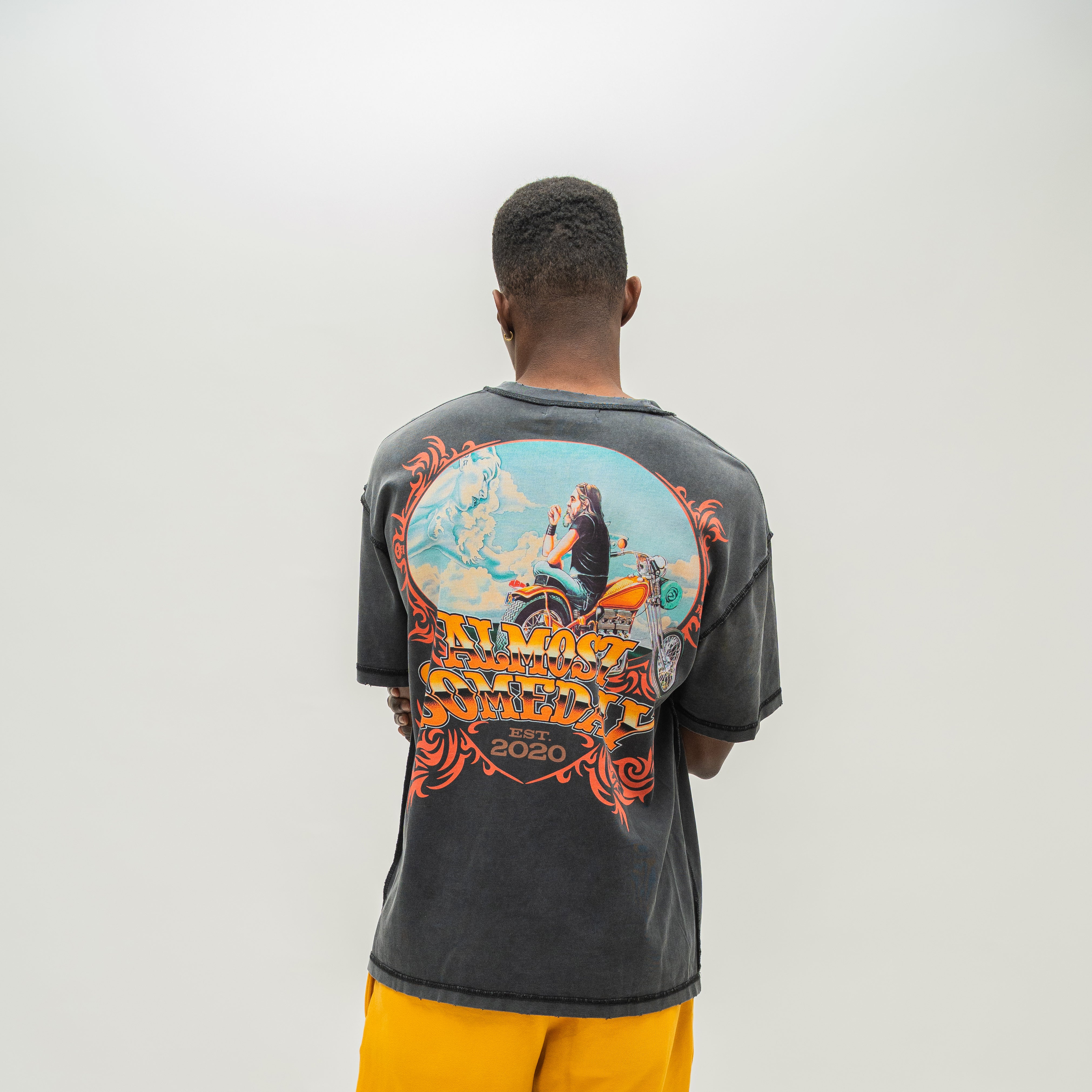 A man viewed from behind wearing a faded black mineral-washed t-shirt with a large colorful graphic on the back, paired with yellow sweatshorts, against a plain light gray background.