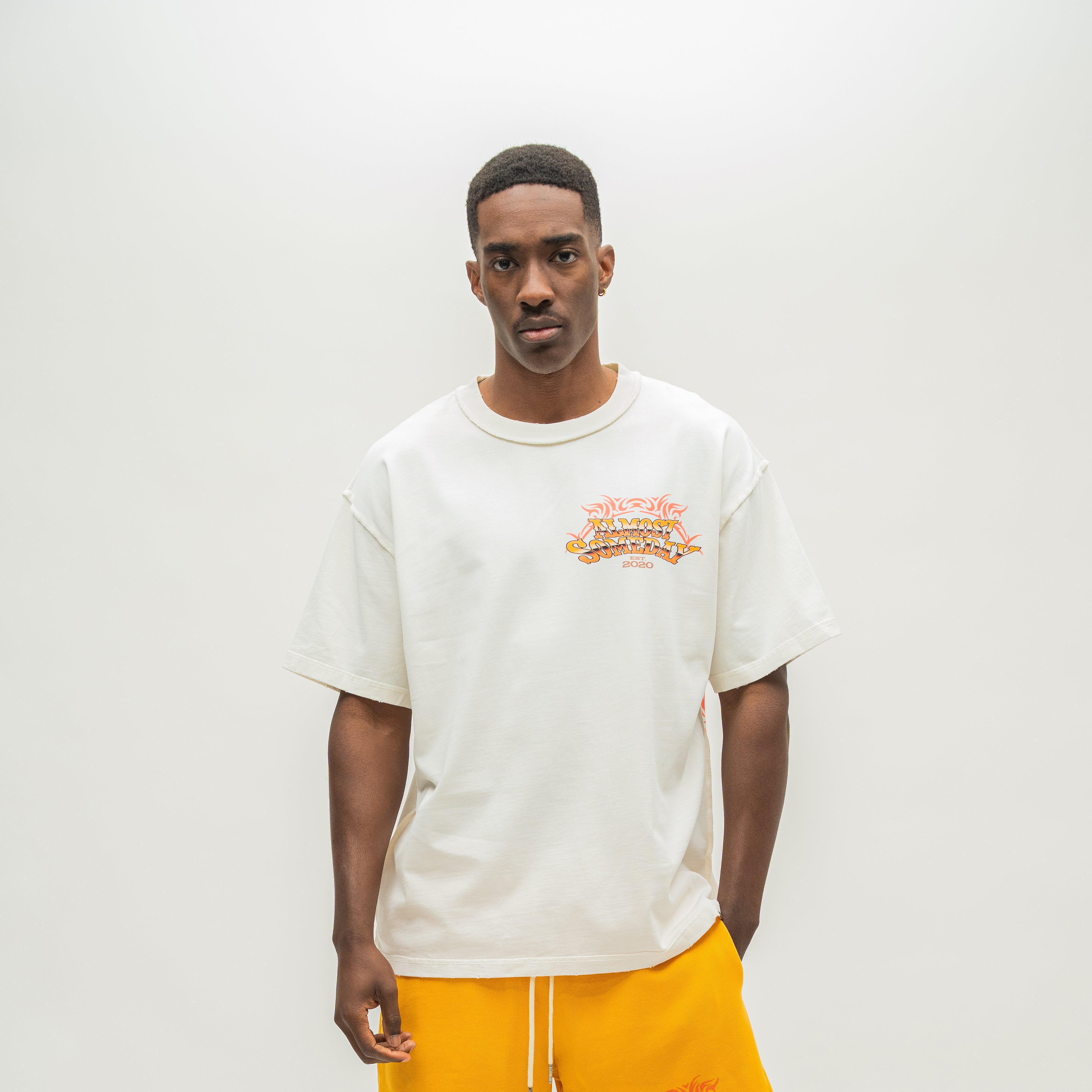 A man wearing a cream-colored short-sleeve t-shirt with a colorful graphic on the left chest, paired with bright yellow sweatpants, standing against a plain light gray background.
