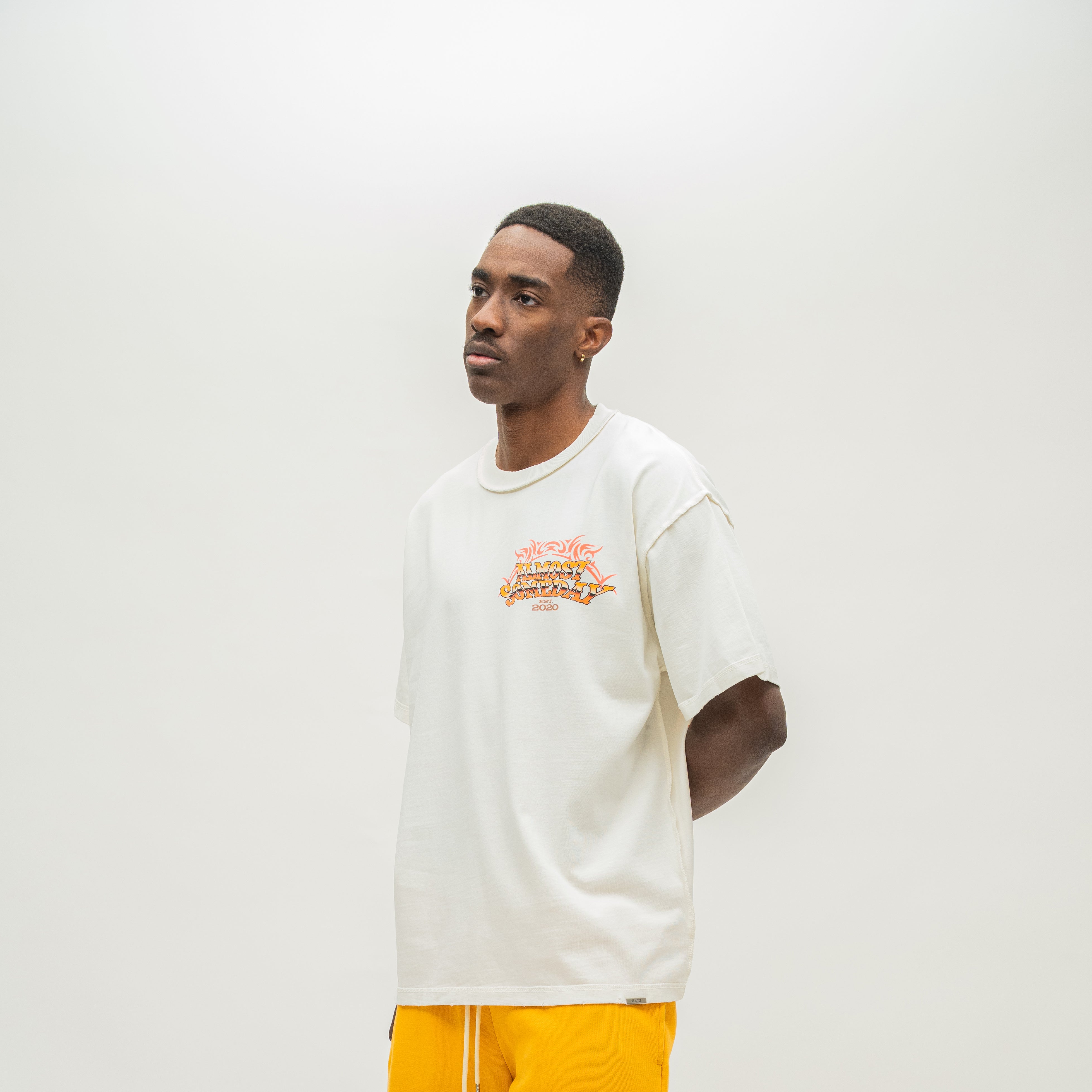 A man wearing a cream-colored t-shirt with a colorful graphic on the chest and bright yellow pants, standing against a plain light gray background.