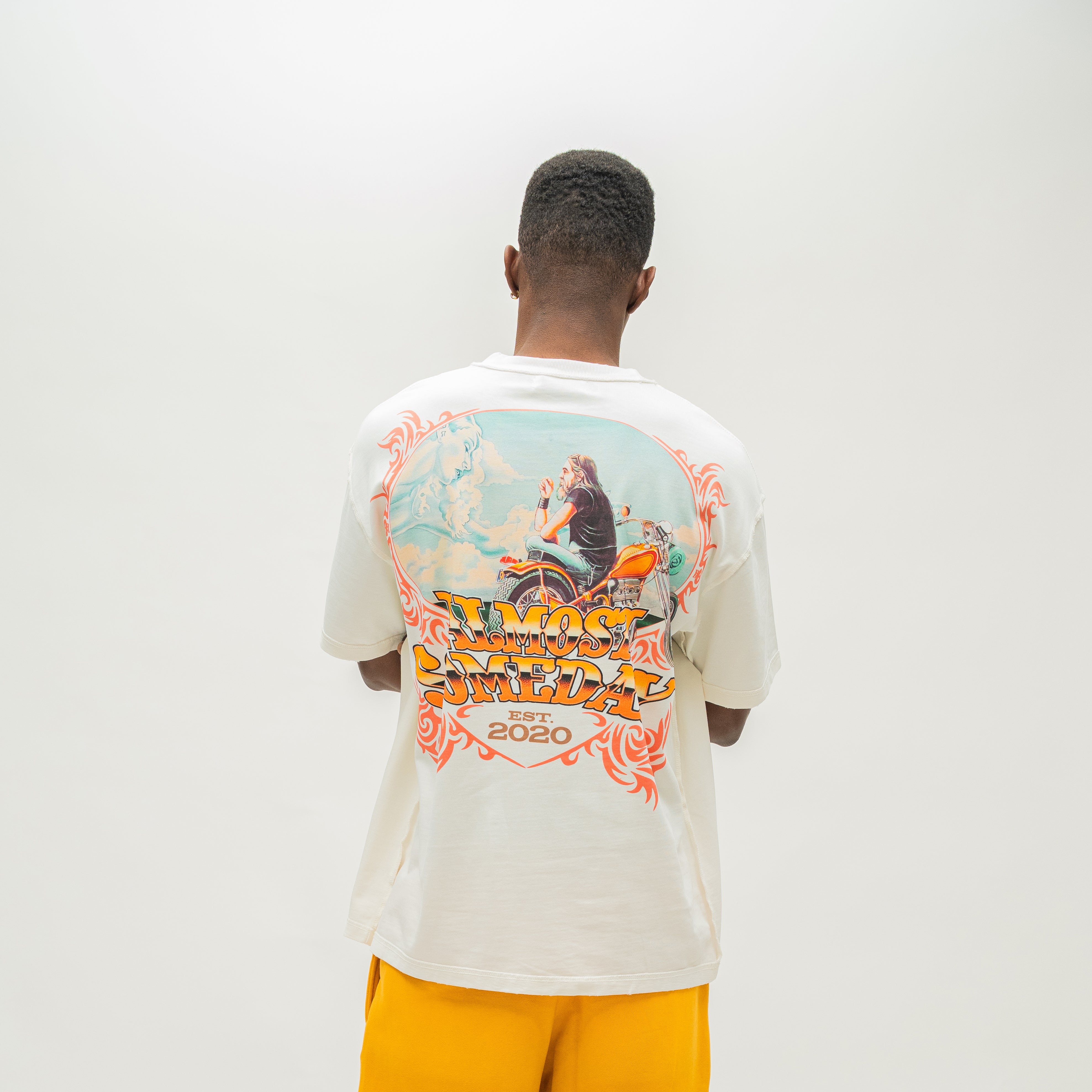 A man viewed from behind wearing a cream-colored graphic t-shirt with a large back print and yellow pants, set against a plain light gray studio background.