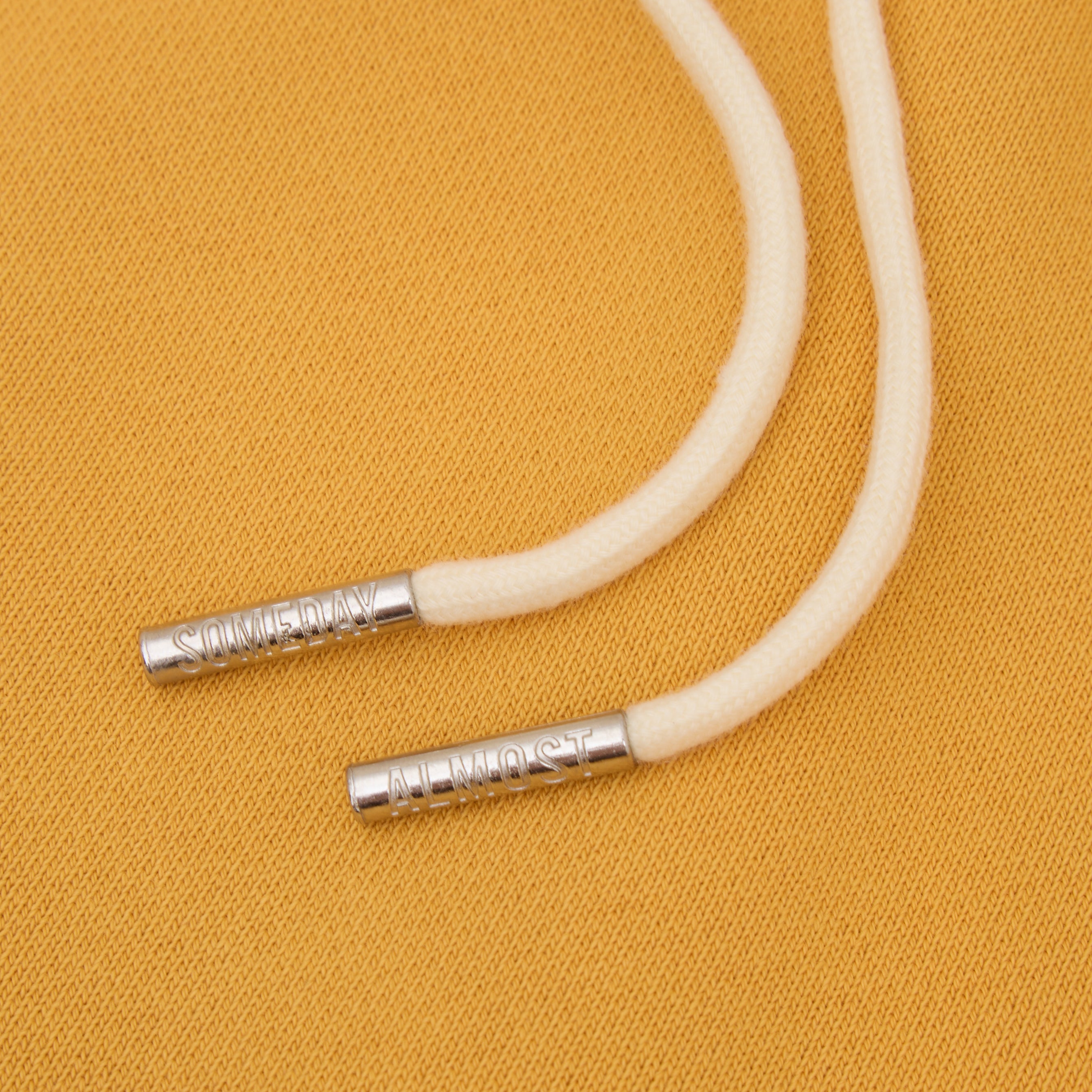 Beige cord with ALMOST SOMEDAY metal ends on a mustard yellow background