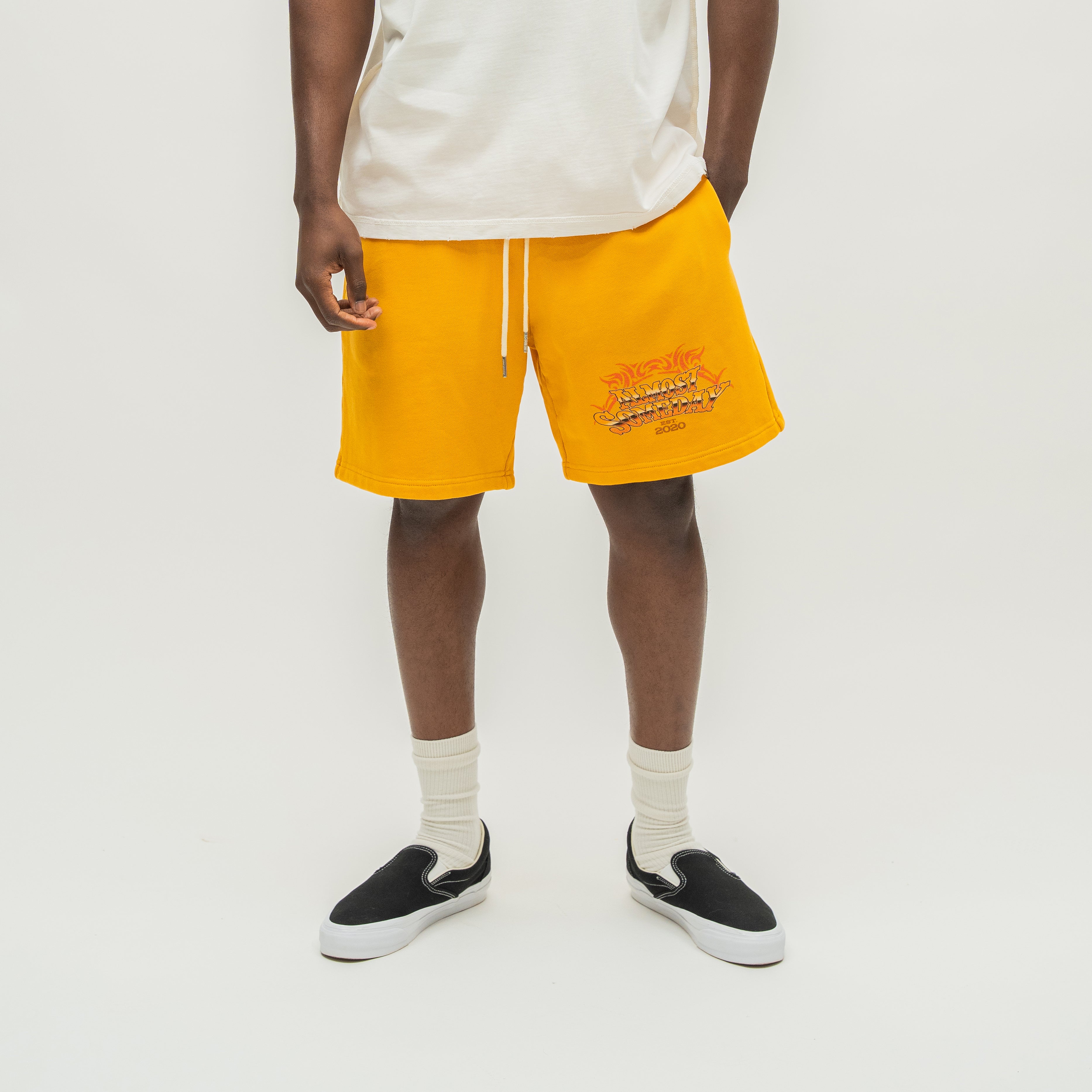 A model wearing bright yellow sweat shorts with a graphic print on the left leg, paired with a white t-shirt, off-white socks, and black slip-on sneakers, shot against a light gray studio background.
