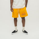 A model wearing bright yellow sweat shorts with a graphic print on the left leg, paired with a white t-shirt, off-white socks, and black slip-on sneakers, shot against a light gray studio background.