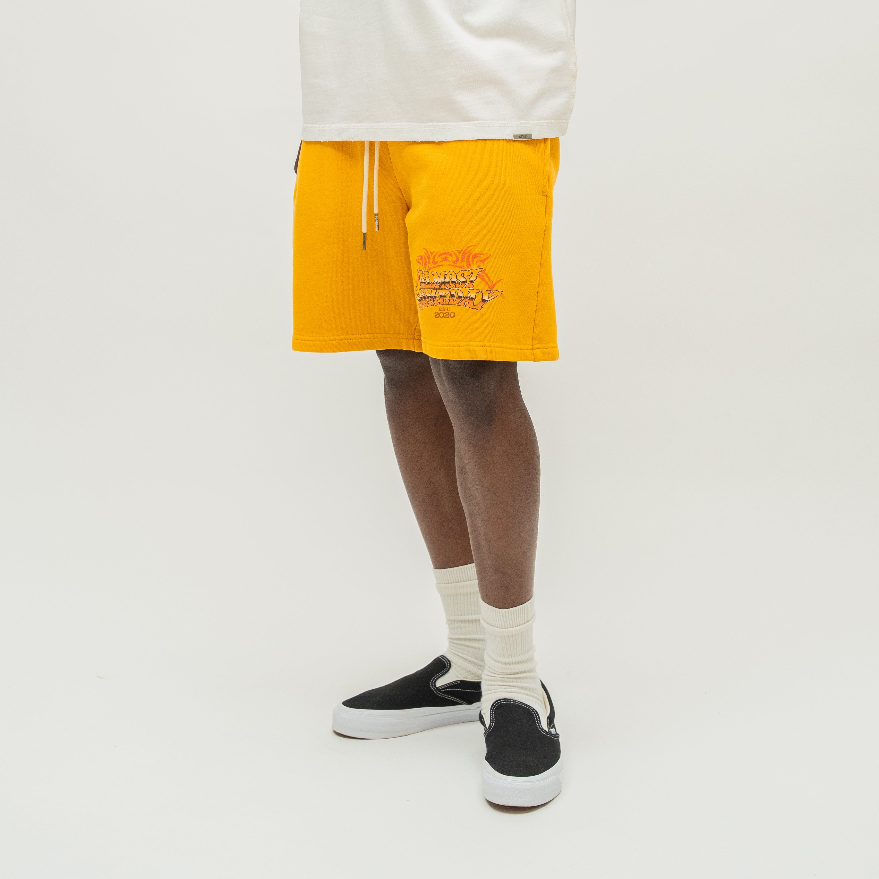 A person wearing bright yellow shorts with a small orange graphic print on the left leg, styled with a white t-shirt, white socks, and black slip-on sneakers against a light gray studio background.