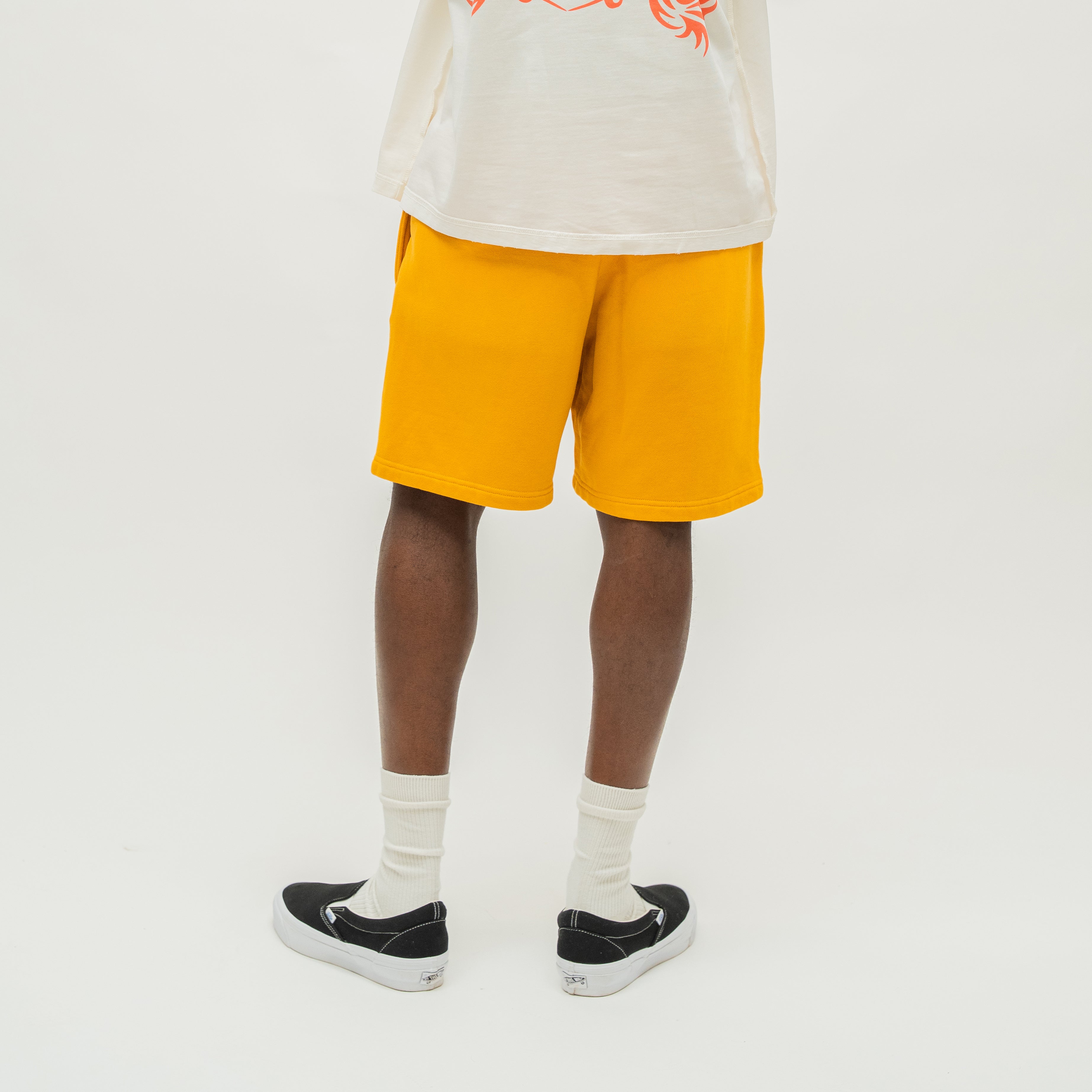 A person shown from the waist down wearing bright yellow shorts, a white t-shirt, white socks, and black slip-on sneakers against a plain white studio background.
