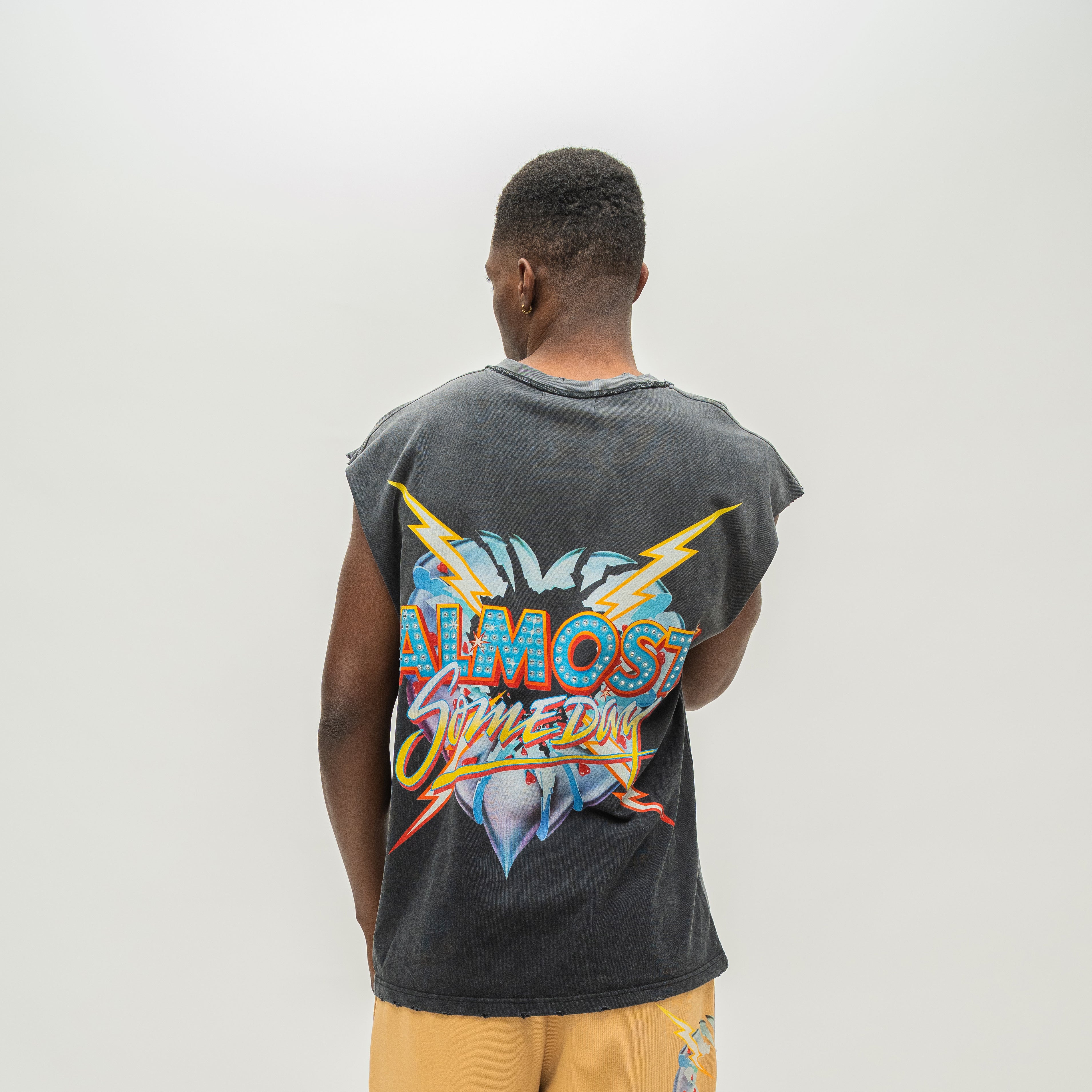 A man viewed from behind wearing a faded black sleeveless t-shirt with a large colorful graphic on the back reading 'ALMOST SOMEDAY', paired with yellow shorts, against a plain light gray background.