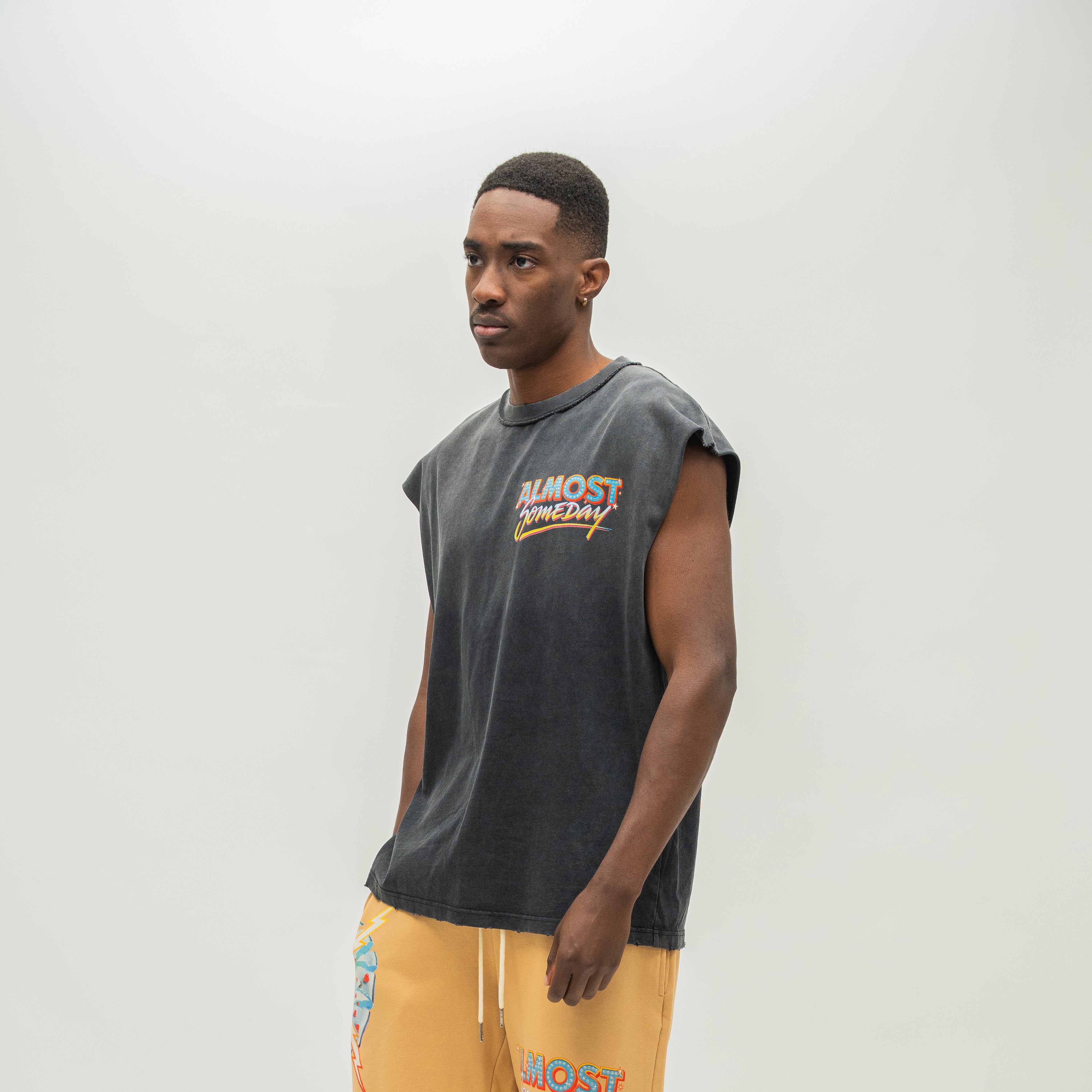 A man wearing a faded black sleeveless t-shirt with a colorful 'ALMOST SOMEDAY' graphic on the chest, paired with yellow sweatshorts, standing against a light gray studio background.