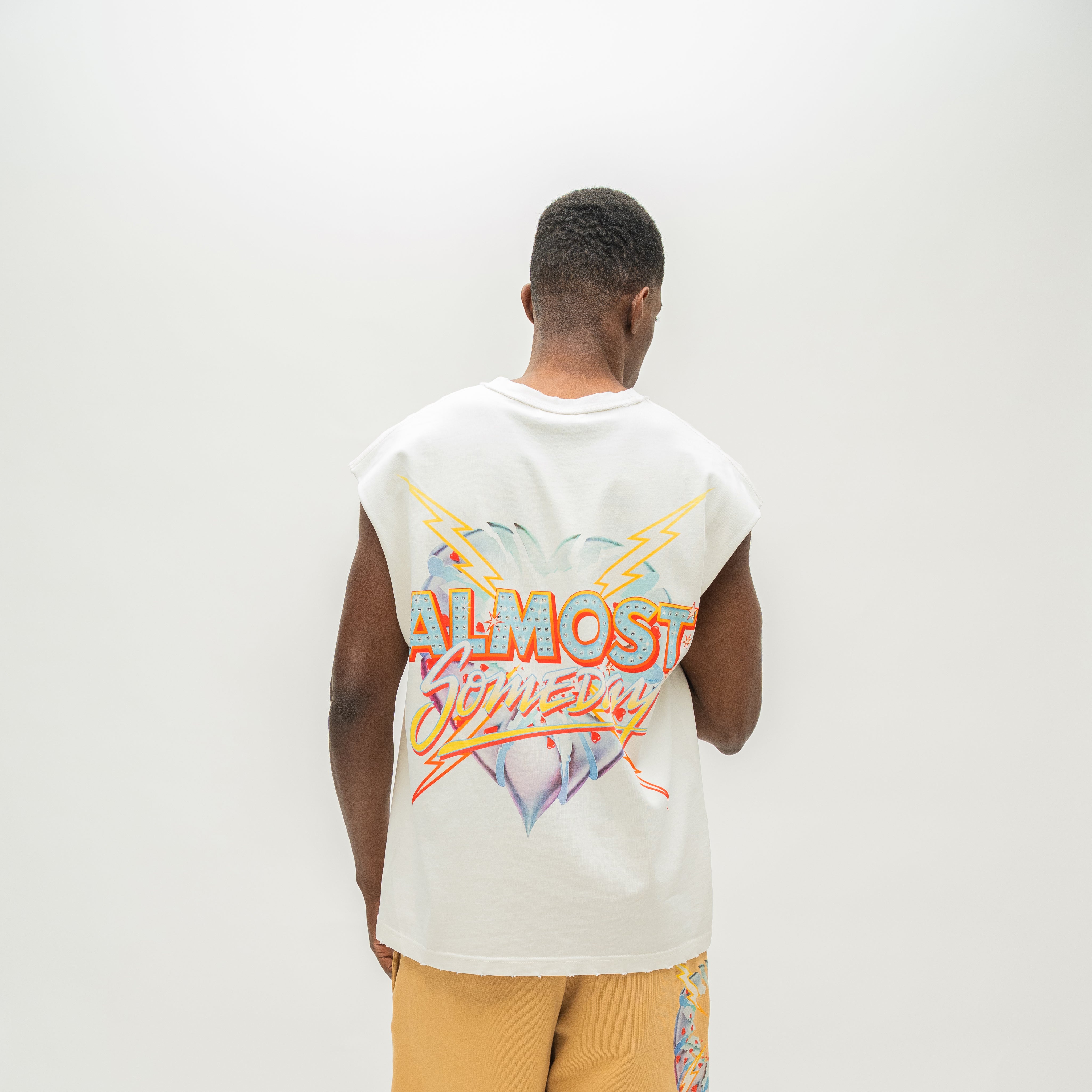 A man viewed from behind wearing a sleeveless white t-shirt with a colorful 'ALMOST SOMEDAY' graphic on the back, paired with yellow shorts, against a plain white studio background.