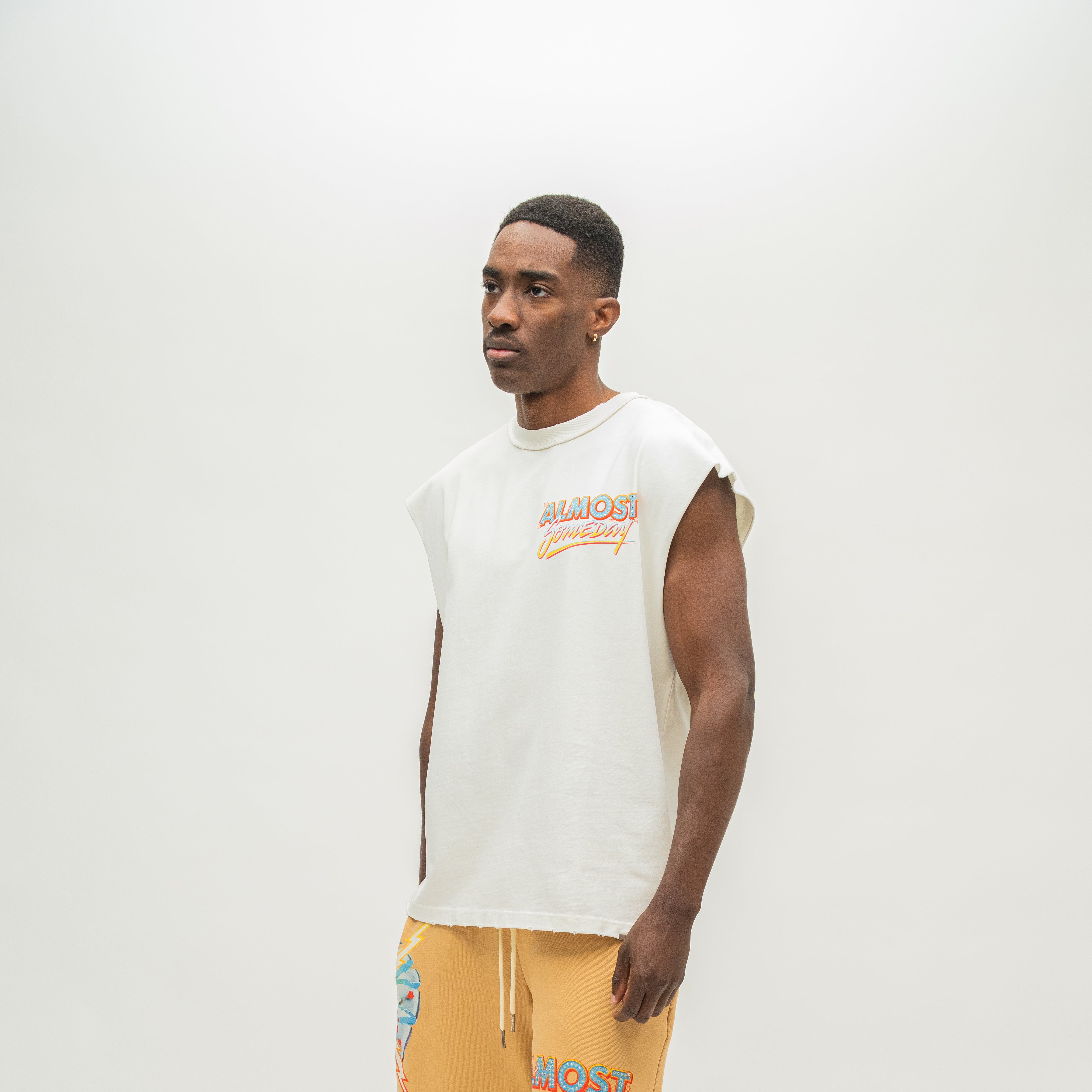 A man wearing a sleeveless white t-shirt with a colorful 'ALMOST SOMEDAY' graphic on the chest, paired with mustard yellow sweatshorts, photographed against a plain light gray studio background.