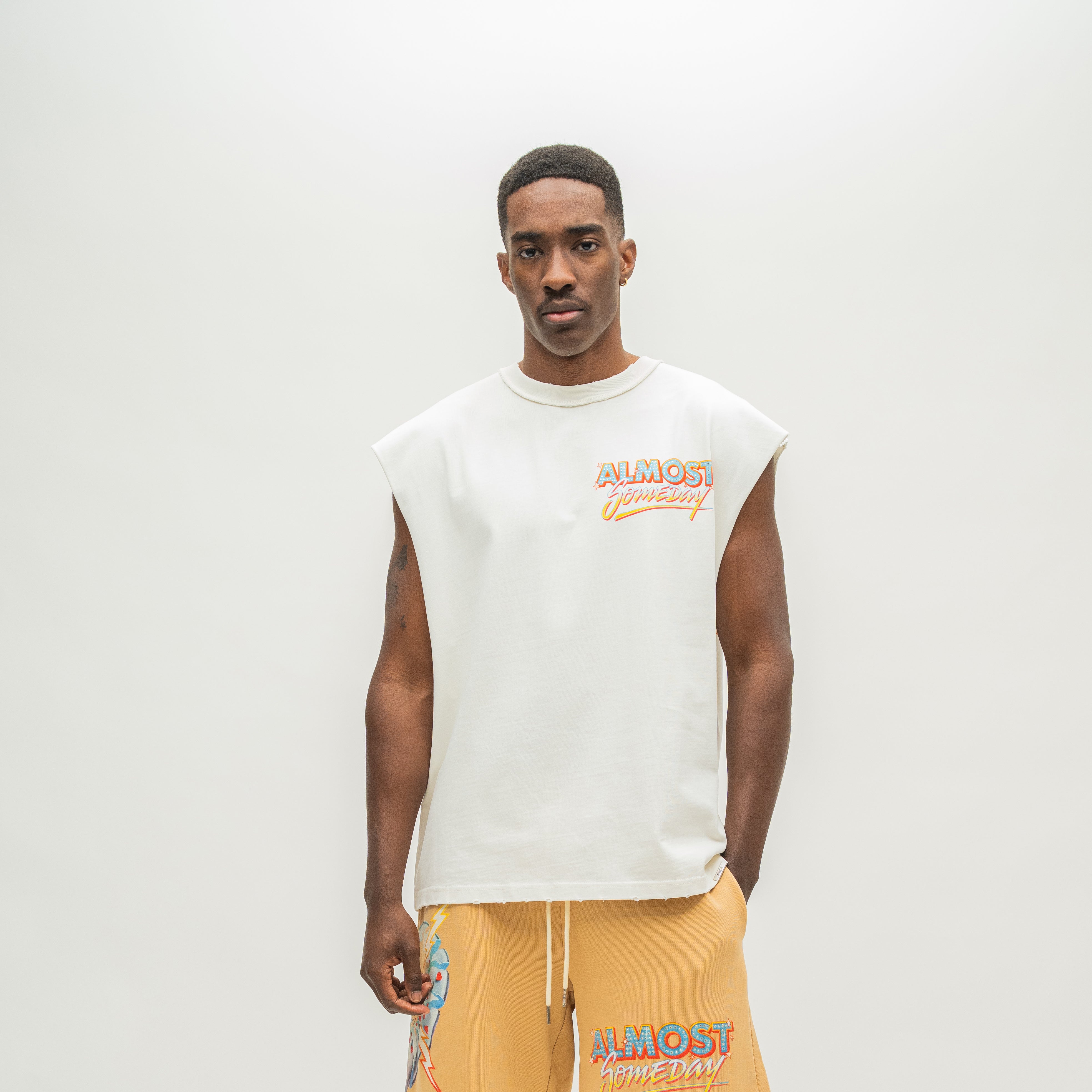 A man modeling a sleeveless white t-shirt with a colorful 'ALMOST SOMEDAY' logo on the chest, paired with mustard yellow sweatshorts against a plain light gray background.