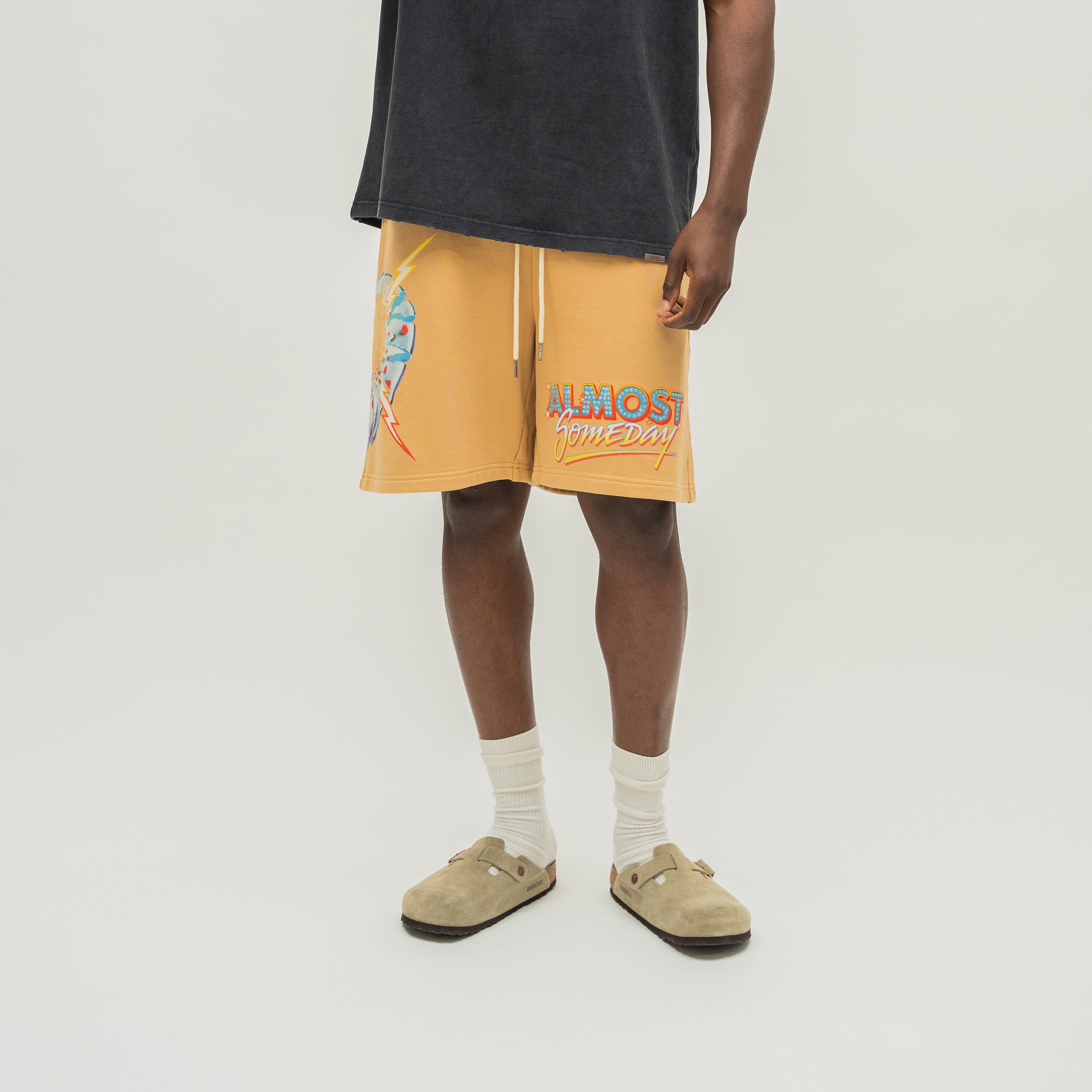 A model wearing yellow-orange drawstring shorts with a graphic print and the text 'ALMOST SOMEDAY' on the left leg, styled with a black t-shirt, white socks, and beige slip-on shoes against a light gray studio background.