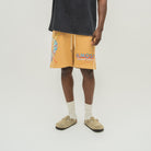 A model wearing yellow-orange drawstring shorts with a graphic print and the text 'ALMOST SOMEDAY' on the left leg, styled with a black t-shirt, white socks, and beige slip-on shoes against a light gray studio background.