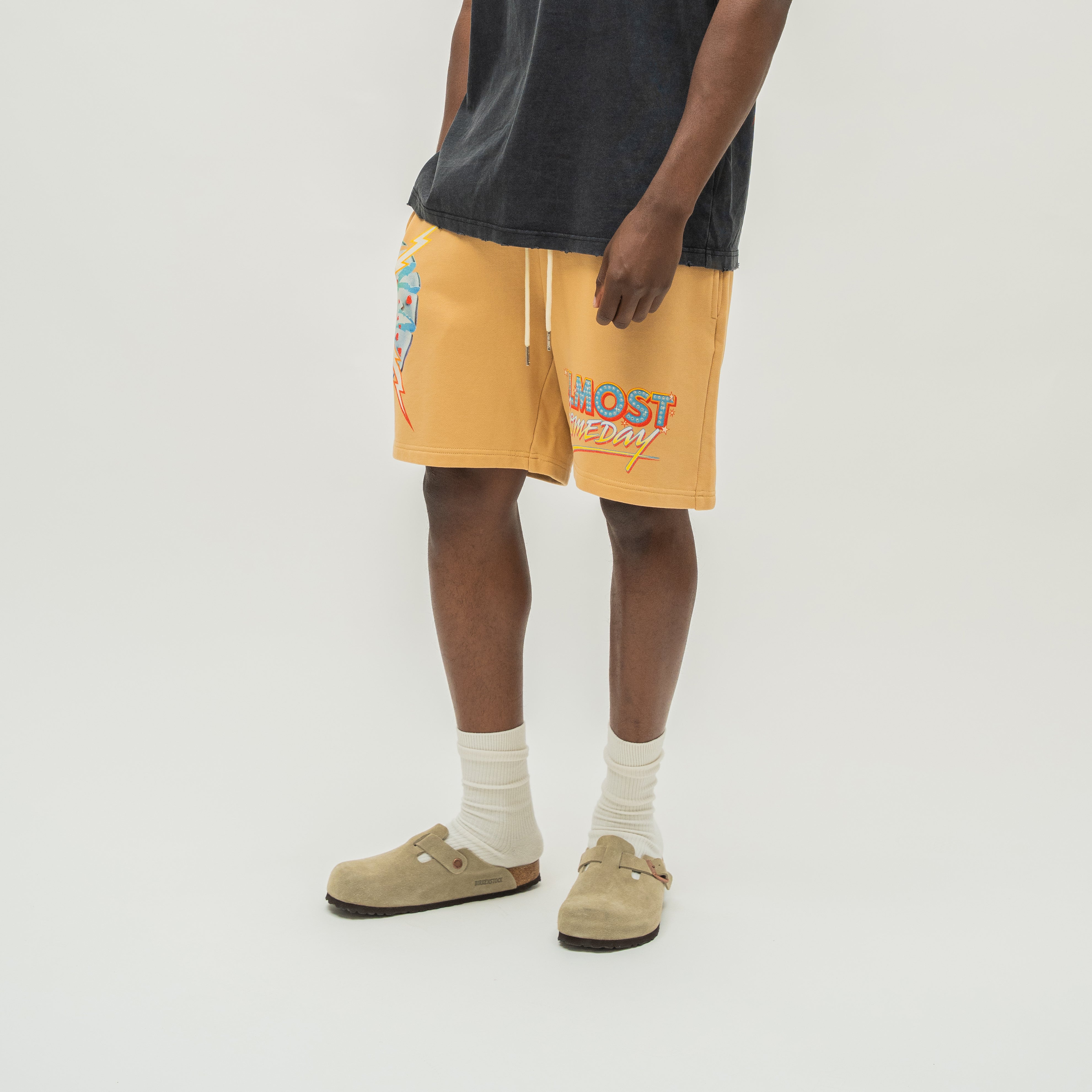 A person wearing mustard yellow drawstring shorts with colorful graphic prints, a faded black t-shirt, white socks, and tan clogs, standing against a light gray studio background.