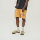 A person wearing mustard yellow drawstring shorts with colorful graphic prints, a faded black t-shirt, white socks, and tan clogs, standing against a light gray studio background.
