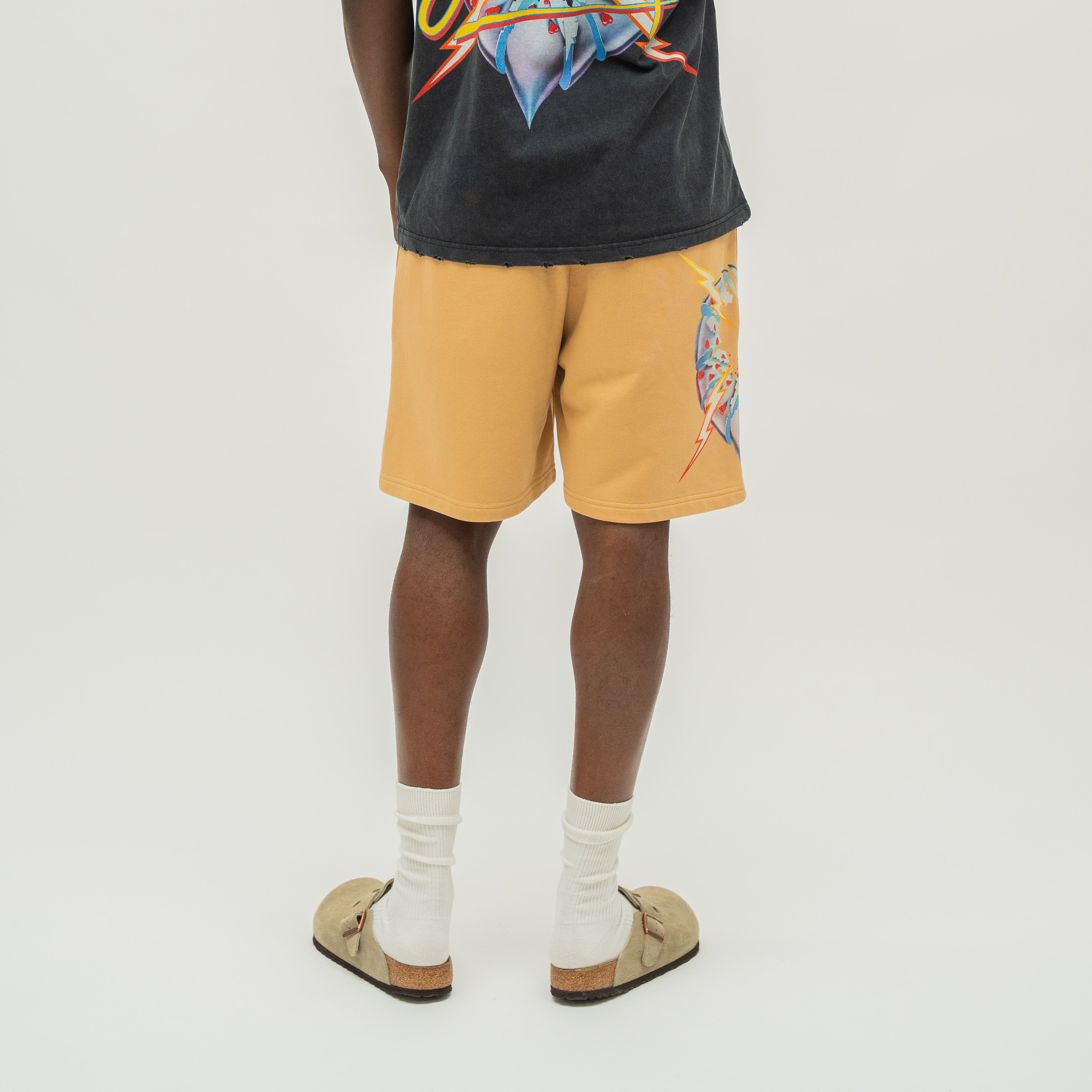 A person shown from the back wearing tan shorts with a colorful graphic print on the left leg, a dark charcoal graphic t-shirt, white socks, and tan loafers, against a plain light gray studio background.