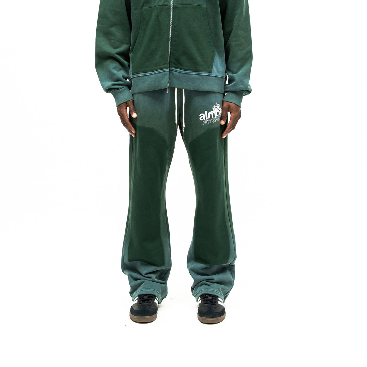 Enemies Flare Bottoms – Green – Front view on model styled with a matching zip hoodie, showing white drawstrings, thigh logo, and dark leg panels