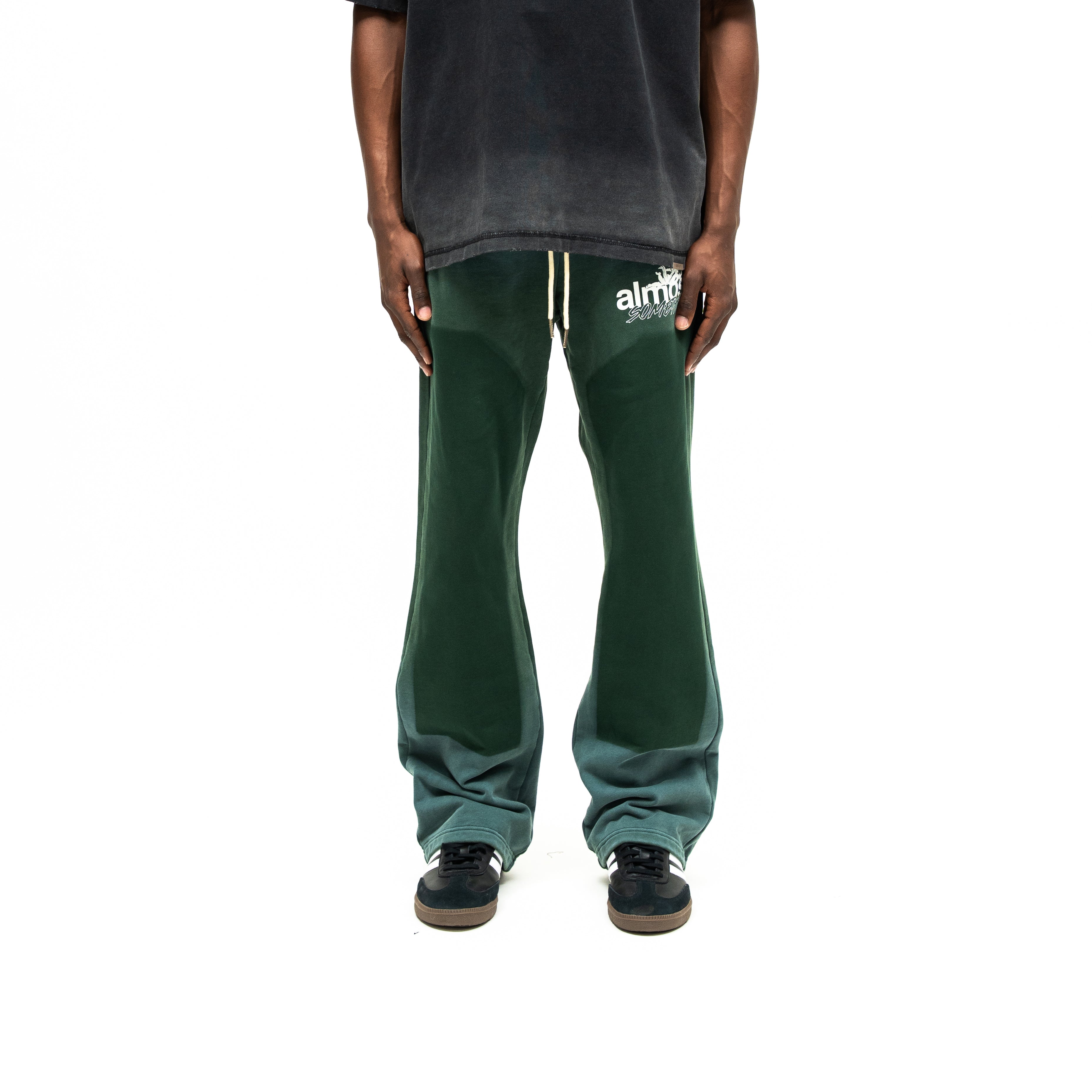 Enemies Flare Bottoms – Green – Front view on model styled with a black tee, showing white thigh logo, drawstrings, and dark leg panels