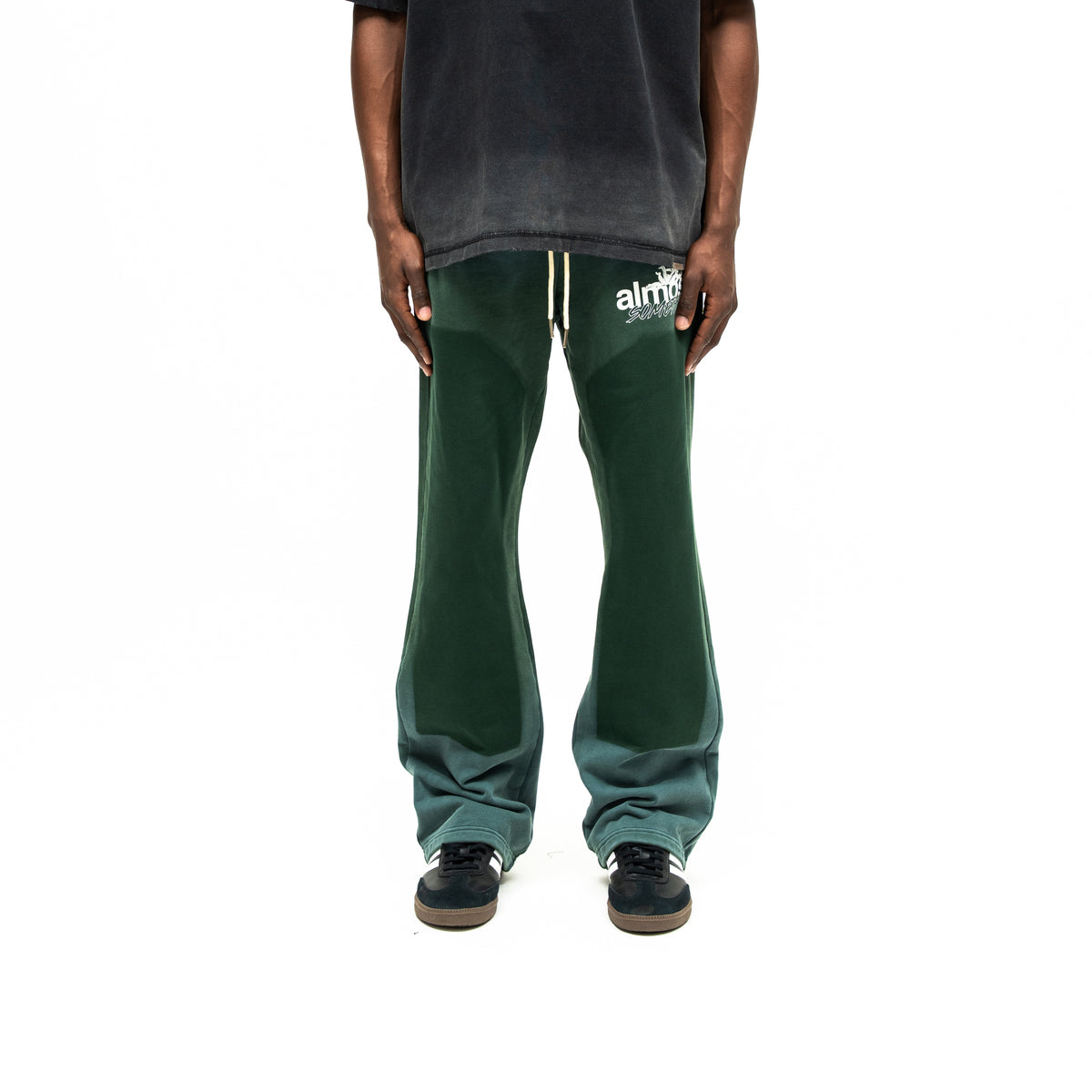 Enemies Flare Bottoms – Green – Front view on model styled with a black tee, showing white thigh logo, drawstrings, and dark leg panels