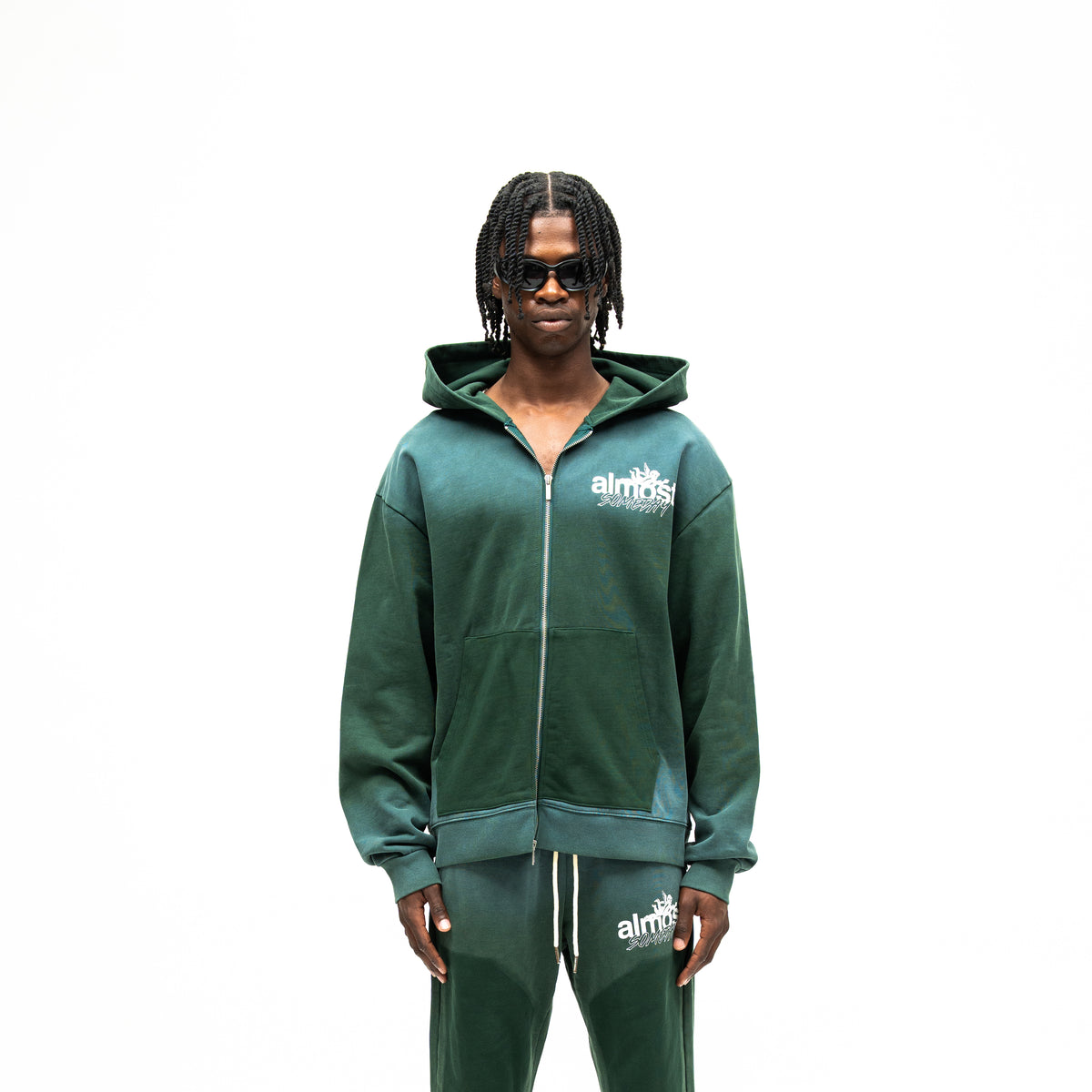 Enemies Zip Hoodie – Green – Front view on model showing full zipper, white chest logo, hood, and dark green front pockets