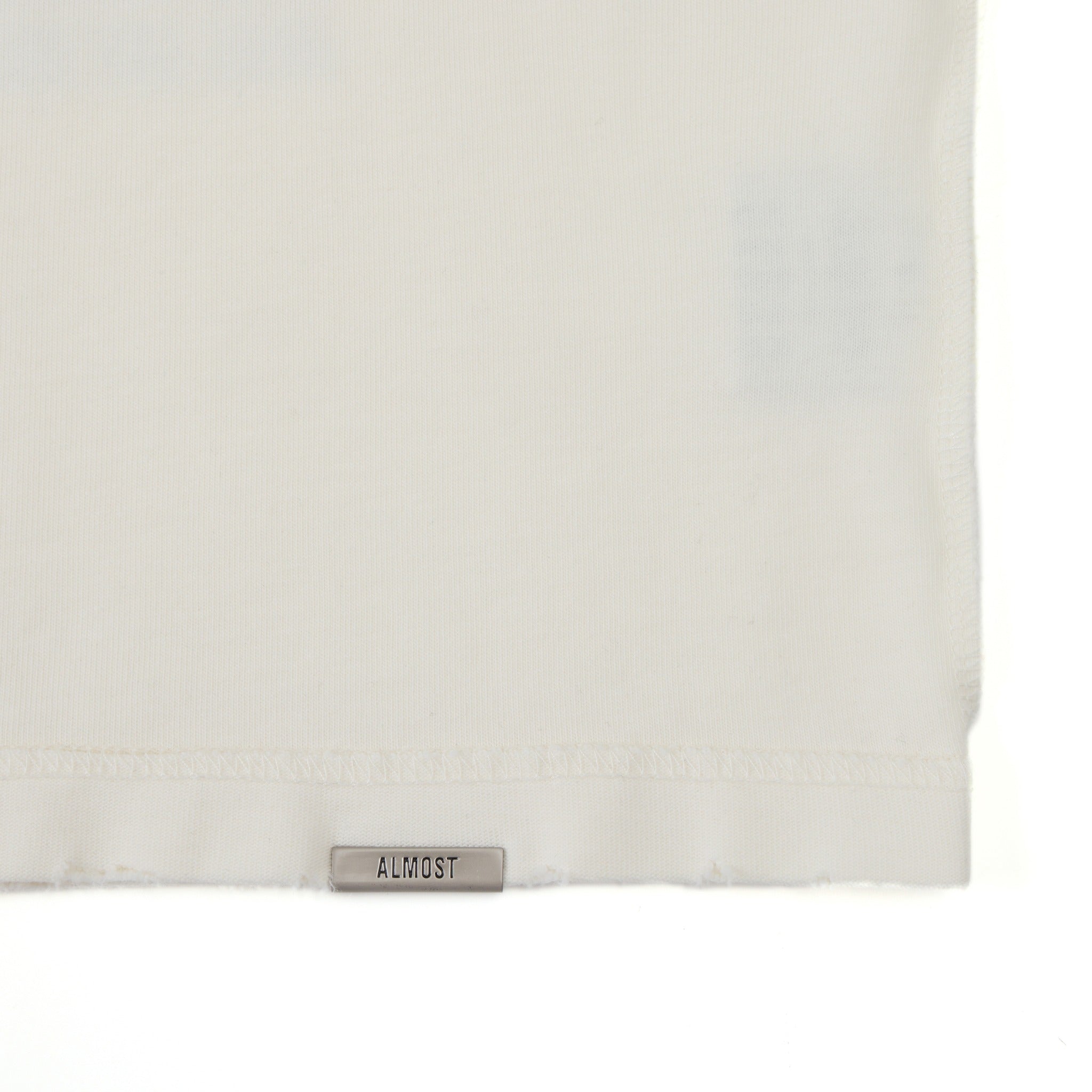 League Tee – Cream – Close-up of metal ALMOST logo at the bottom hem