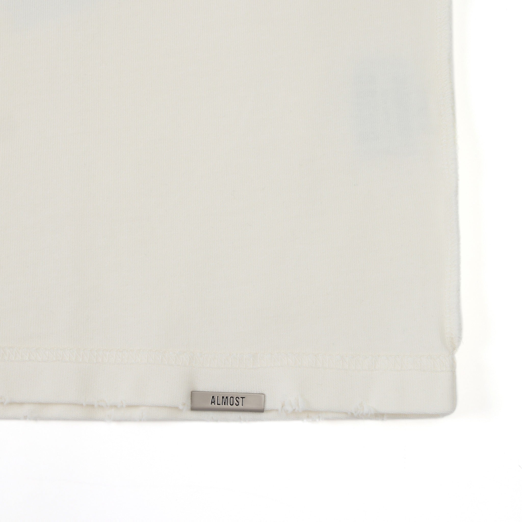 Members Tee – Cream – Close-up of metal ALMOST logo at the bottom hem