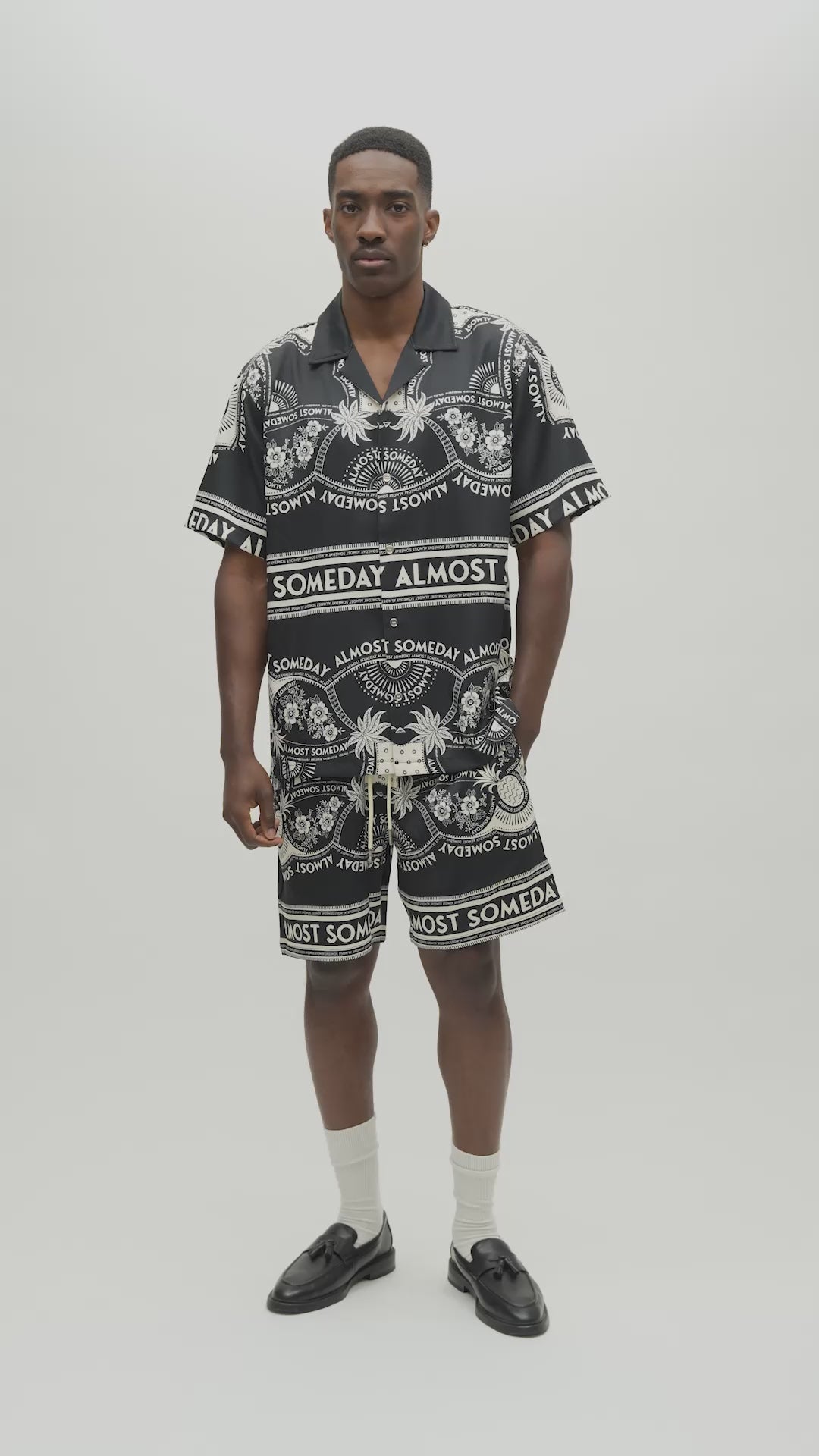 A man modeling a matching black and white patterned short-sleeve button-up shirt and shorts set. The shirt features a bold graphic print with the words 'ALMOST SOMEDAY' and tropical motifs.