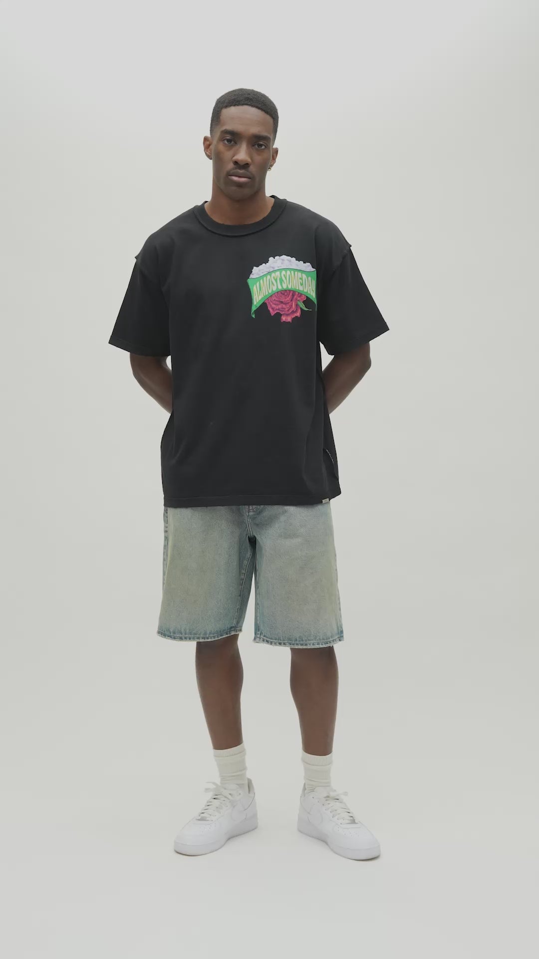 A man wearing a black t-shirt with a colorful 'Almost Someday' graphic on the chest, paired with light blue jeans, standing against a plain light gray background.