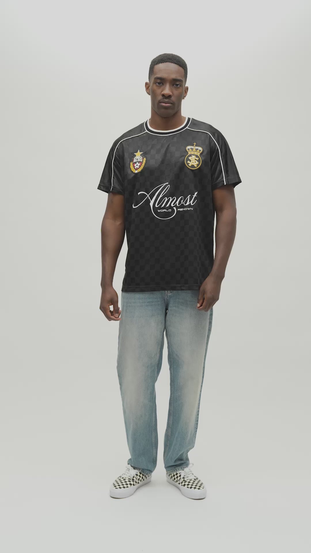 A man wearing a black and dark gray checkered jersey with white piping, a gold crest, and a white 'Almost Someday' logo, paired with light blue jeans, standing against a light gray studio background.