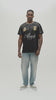A man wearing a black and dark gray checkered jersey with white piping, a gold crest, and a white 'Almost Someday' logo, paired with light blue jeans, standing against a light gray studio background.