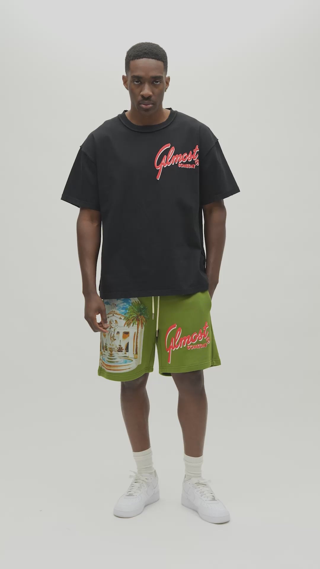A man wearing a black t-shirt with a large graphic print on the back. The graphic features a white building with palm trees under a blue sky, with the red text 'ALMOST SOMEDAY' written across the bottom.