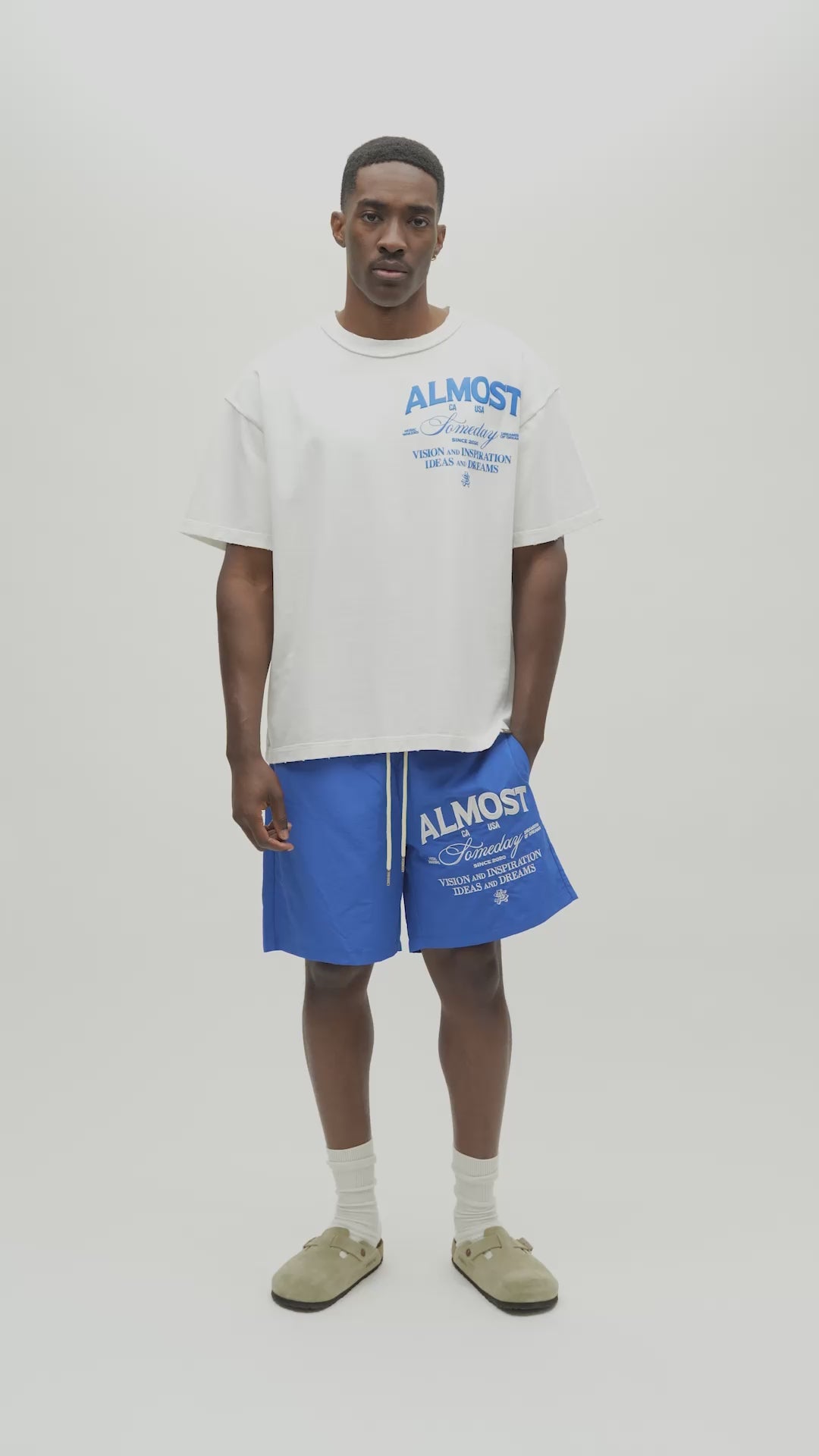 A man wearing an oversized white t-shirt with blue graphic text that reads 'ALMOST SOMEDAY' and blue athletic shorts with matching blue text, standing against a plain off-white studio background.