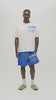 A man wearing an oversized white t-shirt with blue graphic text that reads 'ALMOST SOMEDAY' and blue athletic shorts with matching blue text, standing against a plain off-white studio background.
