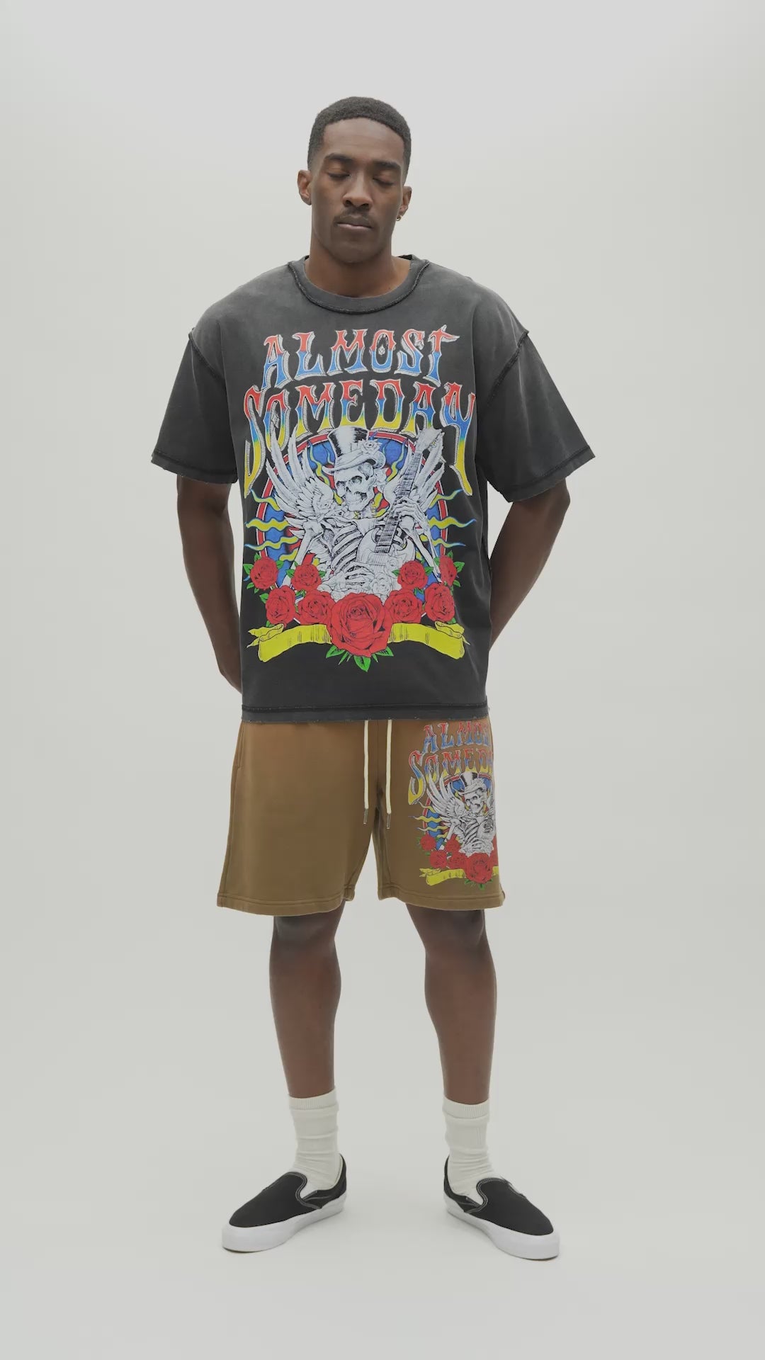 A man wearing a faded black Almost Someday graphic t-shirt with a colorful skeleton and rose design, paired with brown drawstring shorts, standing against a light gray studio background.