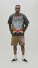 A man wearing a faded black Almost Someday graphic t-shirt with a colorful skeleton and rose design, paired with brown drawstring shorts, standing against a light gray studio background.