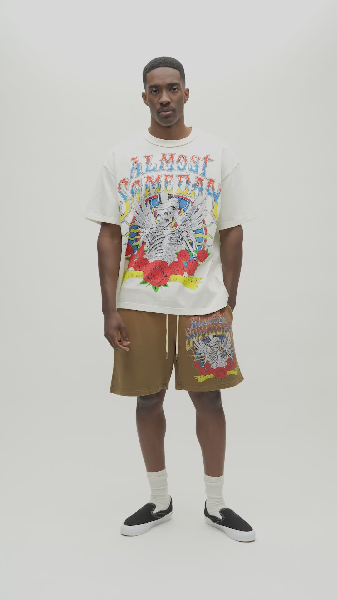 A man wearing a cream-colored graphic t-shirt with a colorful 'Almost Someday' skeleton illustration, paired with brown sweatshorts, against a plain white studio background.