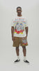 A man wearing a cream-colored graphic t-shirt with a colorful 'Almost Someday' skeleton illustration, paired with brown sweatshorts, against a plain white studio background.