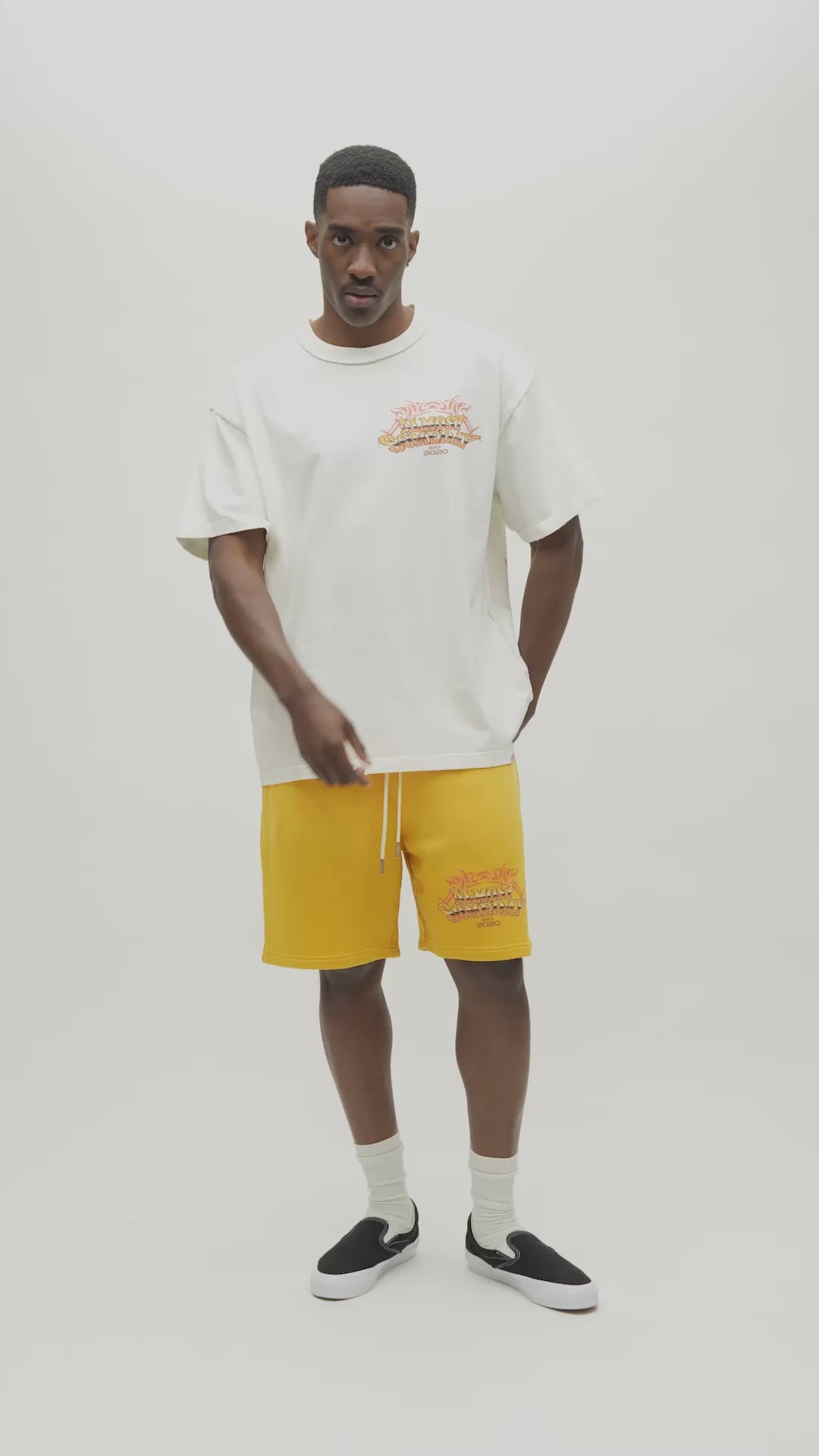 A video of a man wearing a cream-colored graphic t-shirt with a large back print and yellow pants, set against a plain light gray studio background.