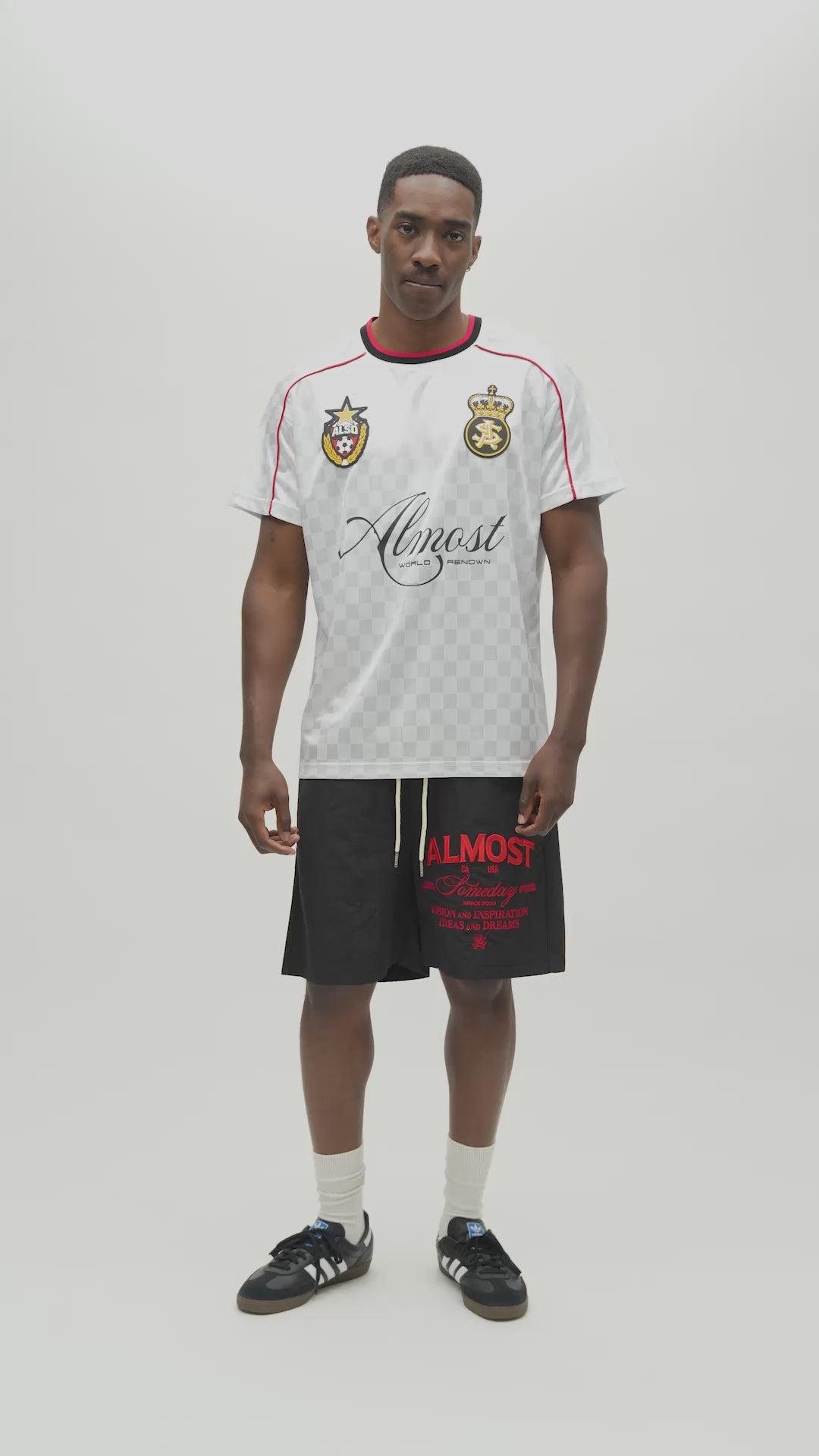 A man modeling a white checkered soccer jersey with red and navy trim, featuring the Almost Someday brand logo on the front, set against a plain light gray background.