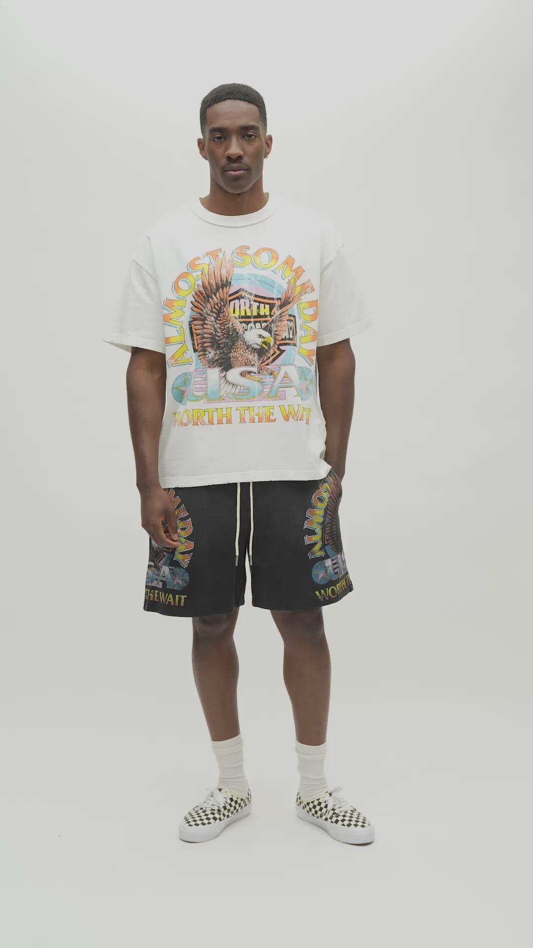 A man wearing a cream-colored graphic t-shirt with a colorful 'Almost Someday USA' design, paired with dark gray shorts, standing against a plain off-white studio background.