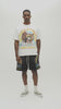A man wearing a cream-colored graphic t-shirt with a colorful 'Almost Someday USA' design, paired with dark gray shorts, standing against a plain off-white studio background.