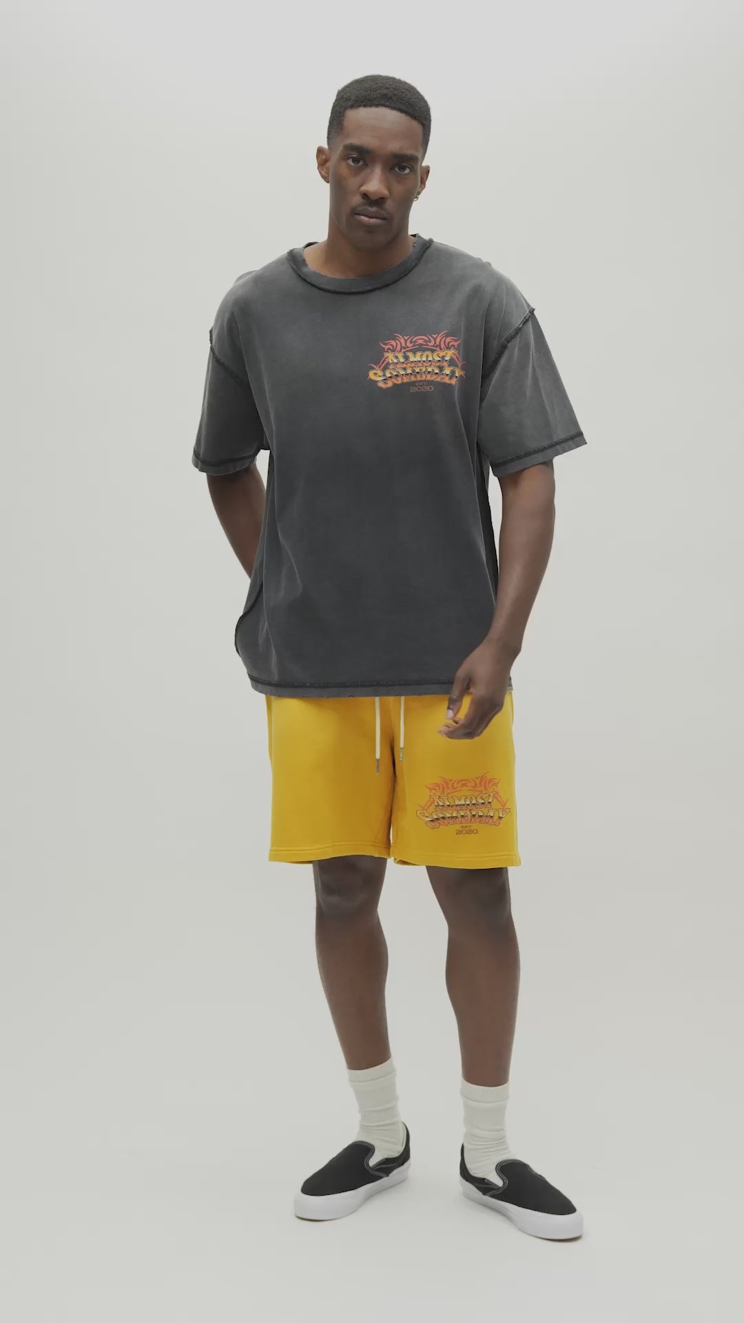 A man wearing a faded black mineral-washed t-shirt with a large colorful graphic on the back, paired with yellow sweatshorts, against a plain light gray background.