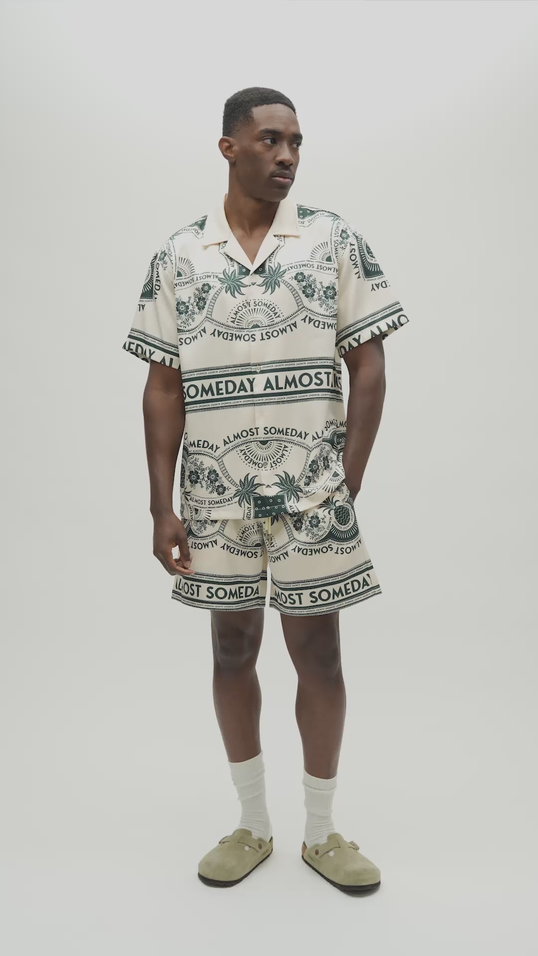 A man wearing a matching short-sleeve camp collar shirt and shorts set with a cream base and dark green graphic print featuring palm trees and the text 'ALMOST SOMEDAY'.