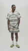 A man wearing a matching short-sleeve camp collar shirt and shorts set with a cream base and dark green graphic print featuring palm trees and the text 'ALMOST SOMEDAY'.