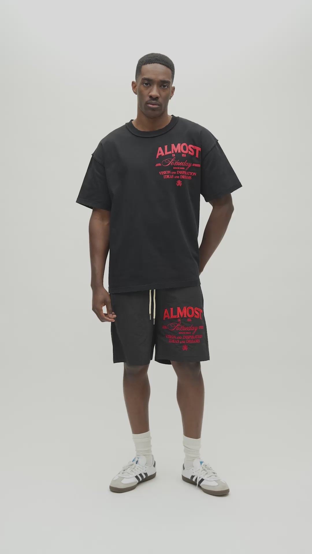 A man viewed from behind wearing a black t-shirt with a large graphic print on the back. The graphic features a white architectural illustration of a cathedral flanked by palm trees, with the words 'ALMOST Someday' in red and white text.