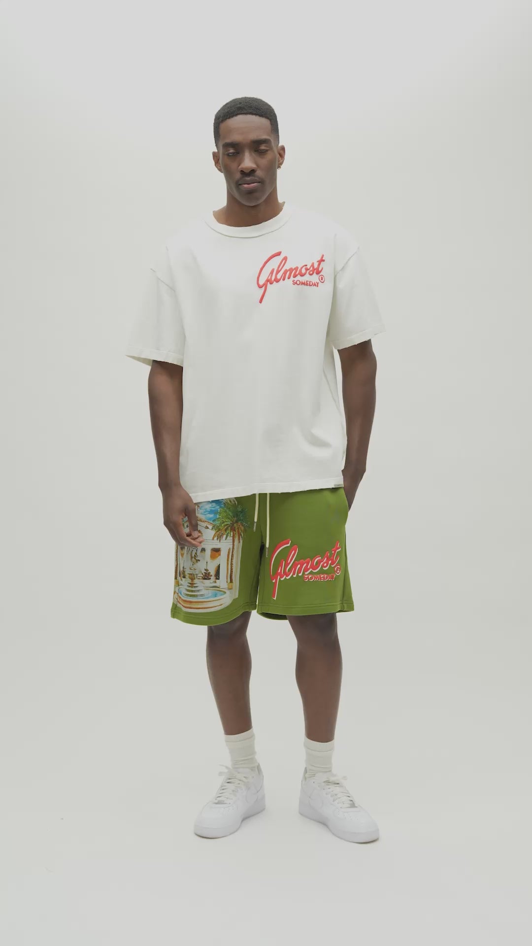A man wearing an off-white t-shirt with a red 'Almost Someday' logo and green shorts with a beach graphic and red logo, standing against a plain light gray background.