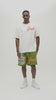 A man wearing an off-white t-shirt with a red 'Almost Someday' logo and green shorts with a beach graphic and red logo, standing against a plain light gray background.